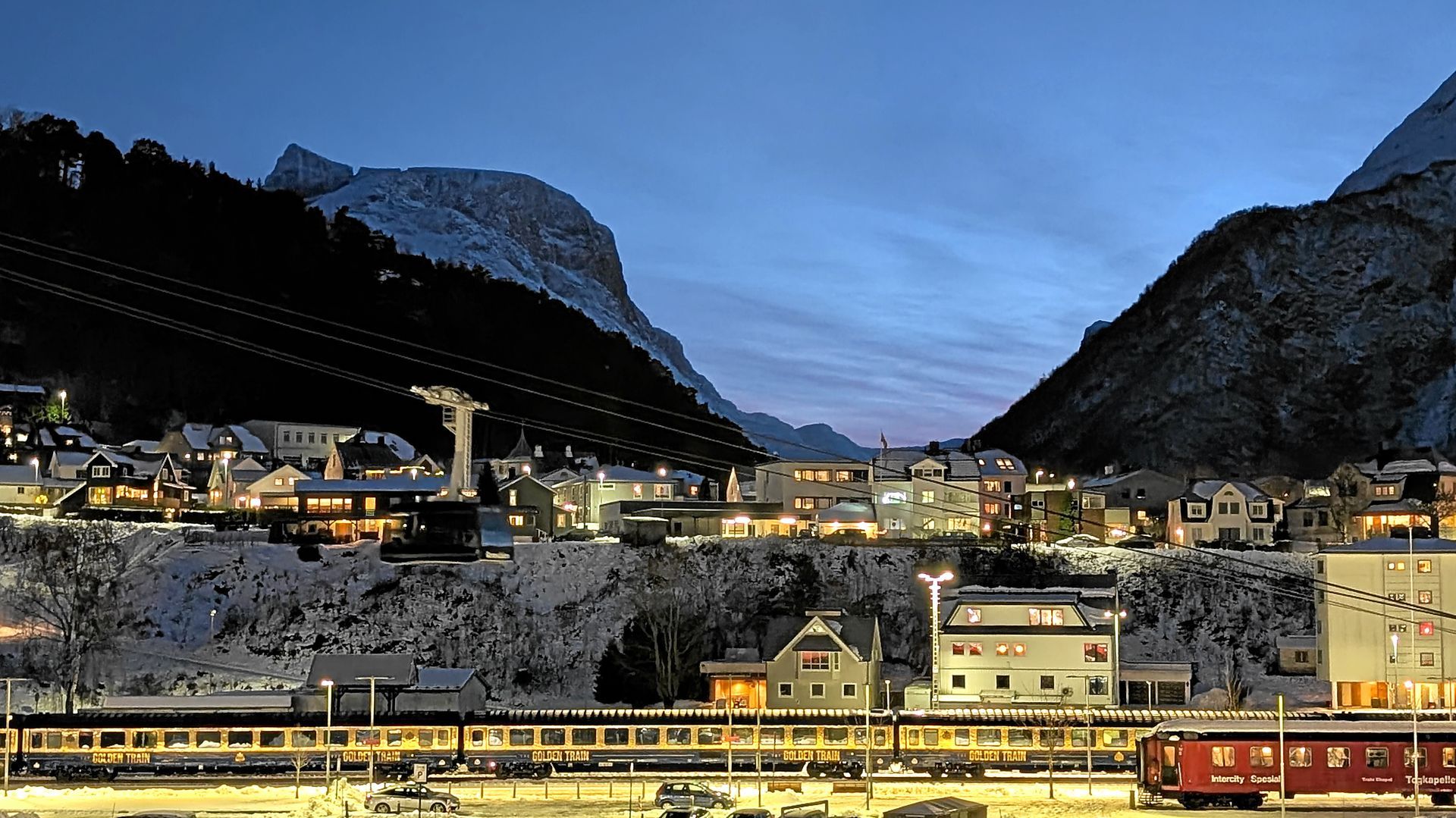 Eine schneebedeckte Stadt, eingebettet zwischen Bergen in der Abenddämmerung. Zug und Seilbahn sind zu sehen.