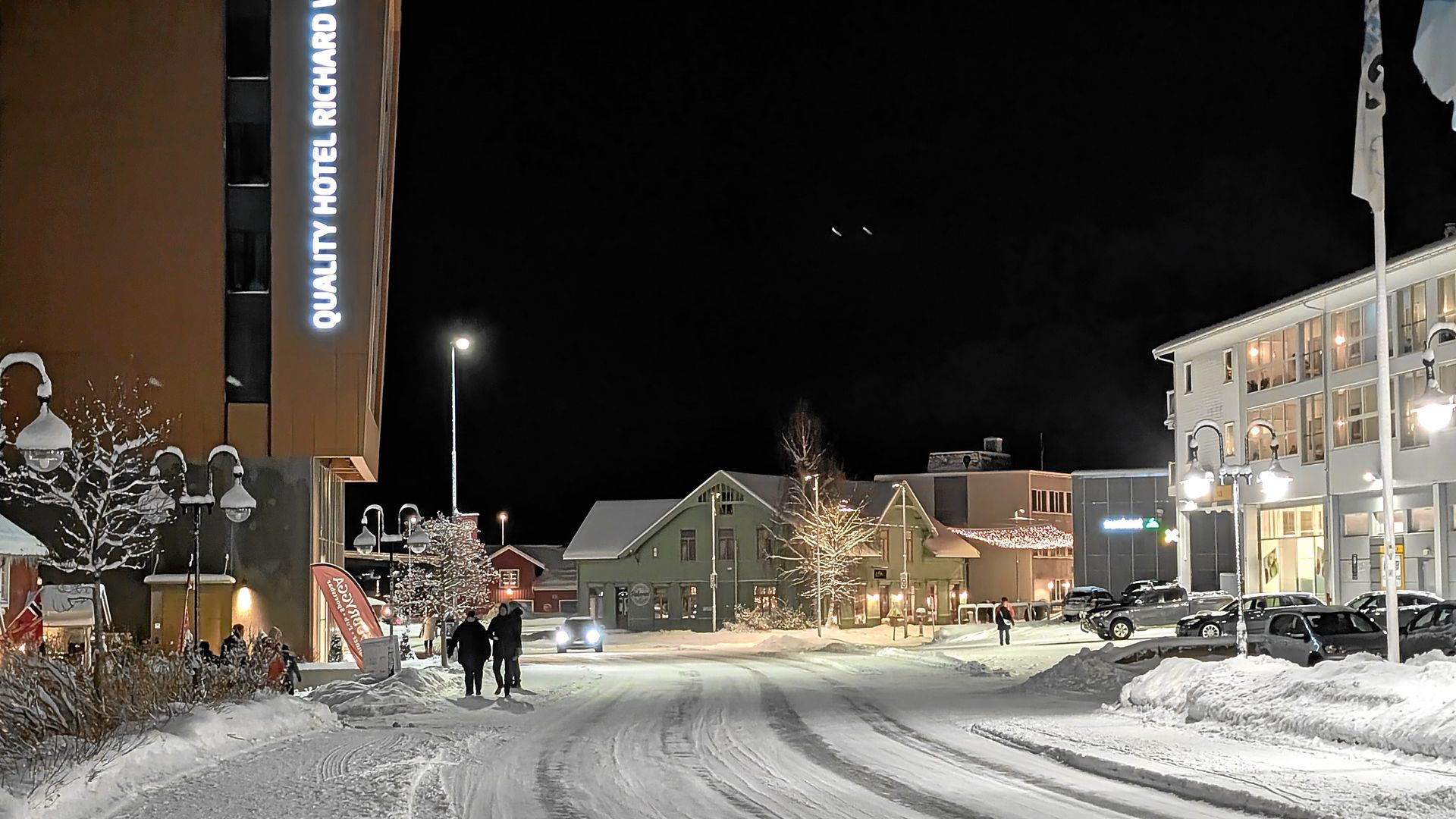 Nachts verschneite Straßenszene mit Hotel und Gebäuden, Weihnachtsbeleuchtung und Passanten.