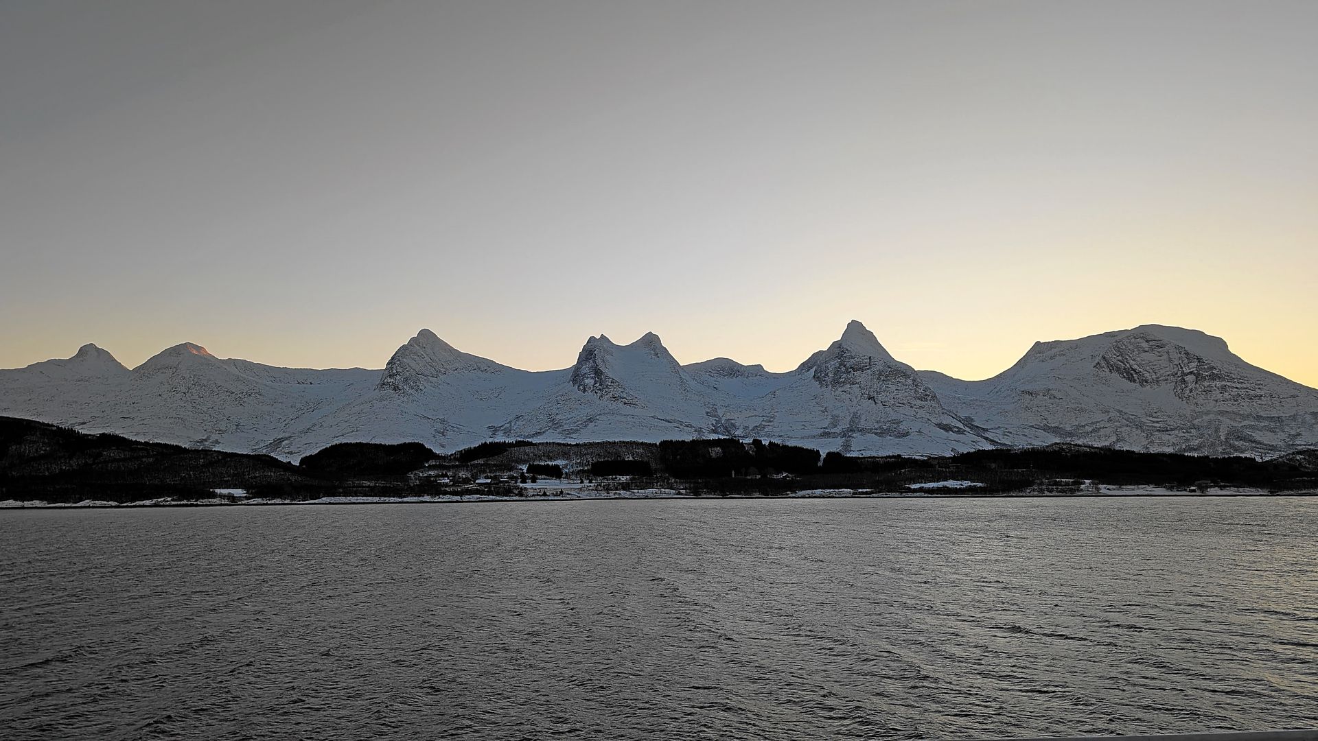Schneebedeckte Berge säumen den Horizont über einem schimmernden Gewässer in der Abenddämmerung.
