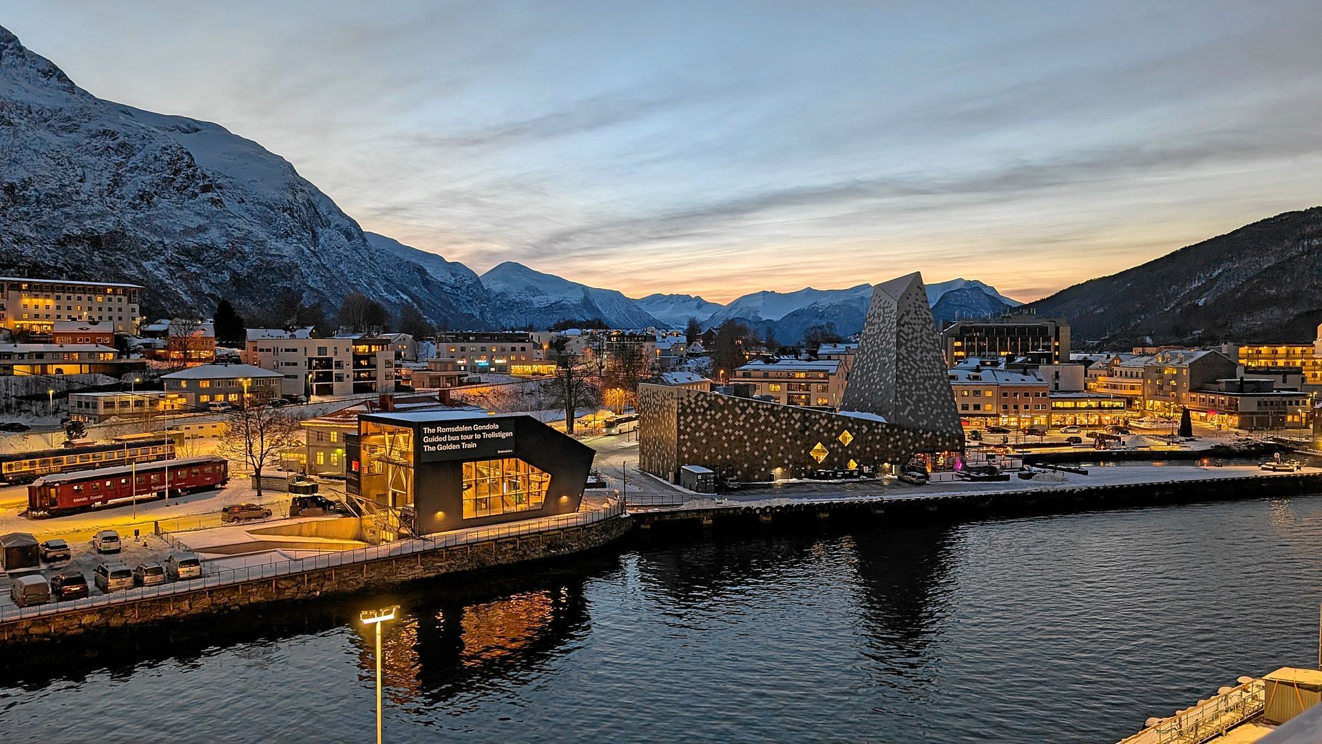 Stadtbild am Wasser mit Bergen in der Abenddämmerung. Gebäude, ein Pier, Schnee und ein bewölkter Himmel.