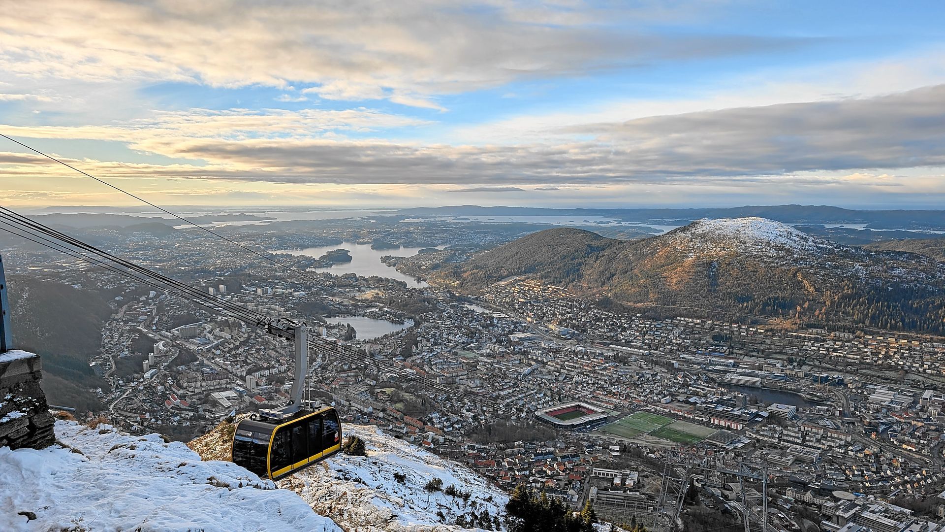 Seilbahnstation auf einem Berg mit Blick auf die schneebedeckte Stadt und die Fjorde unter bewölktem Himmel.