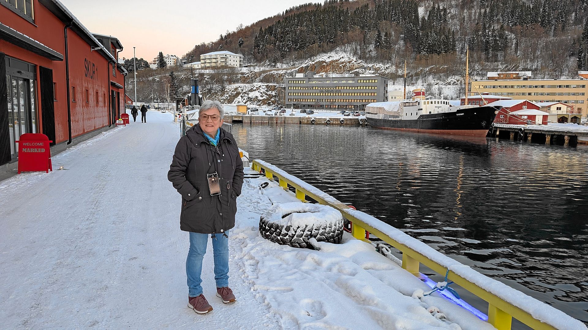 Eine Person steht an einem verschneiten Hafen. Im Hintergrund sind Gebäude und ein Berg zu sehen.