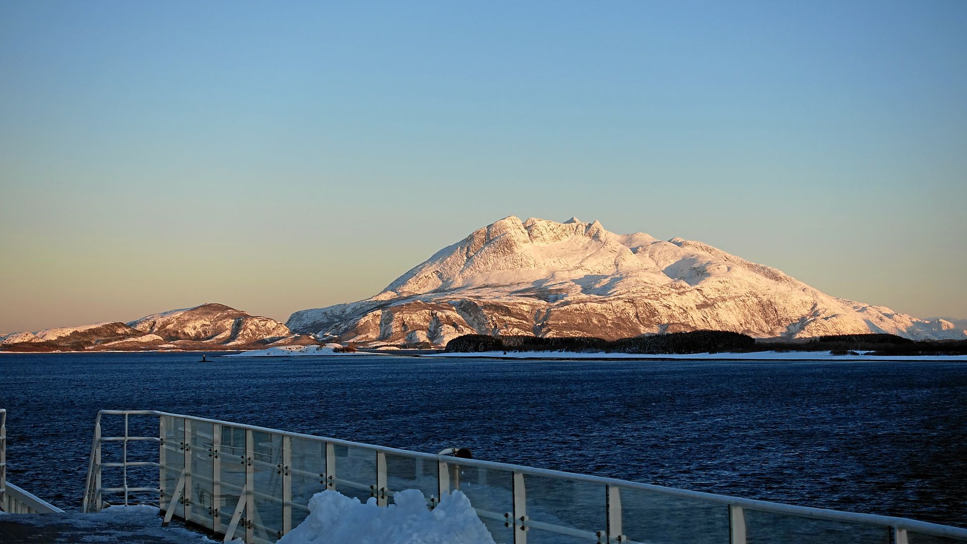 Schneebedeckter Berg über dunkelblauem Wasser, gesehen von einer schneebedeckten Terrasse. Klarer Himmel im Morgengrauen.