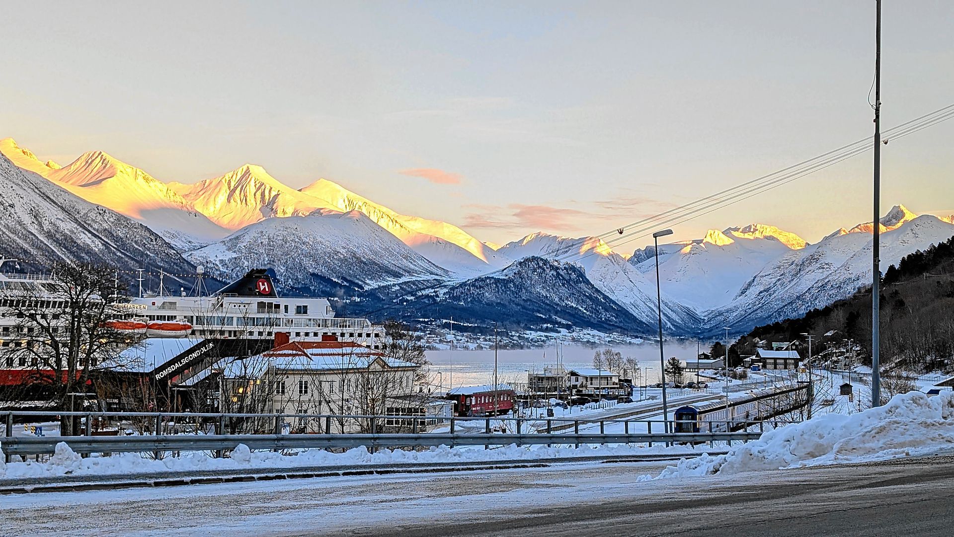Verschneite Bergkette bei Sonnenaufgang, Stadt im Vordergrund mit schneebedeckten Gebäuden, ein Gewässer im Mittelgrund.