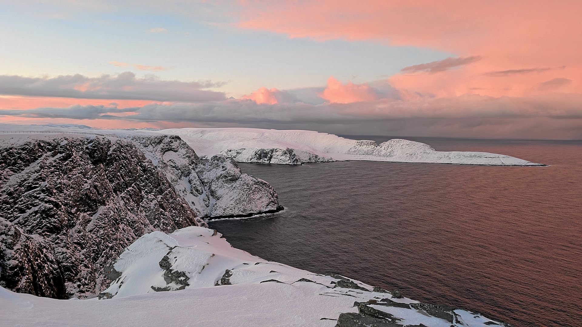 Schneebedeckte Küstenlandschaft bei Sonnenuntergang, rosafarbener und blauer Himmel über dem Meer.