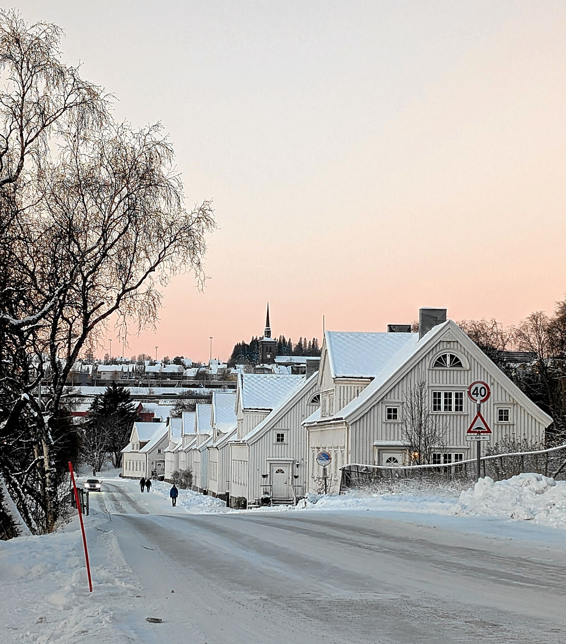 Schneebedeckte Straße mit weißen Häusern in der Abenddämmerung. Zwei Personen gehen die Straße entlang; blassrosa Himmel.