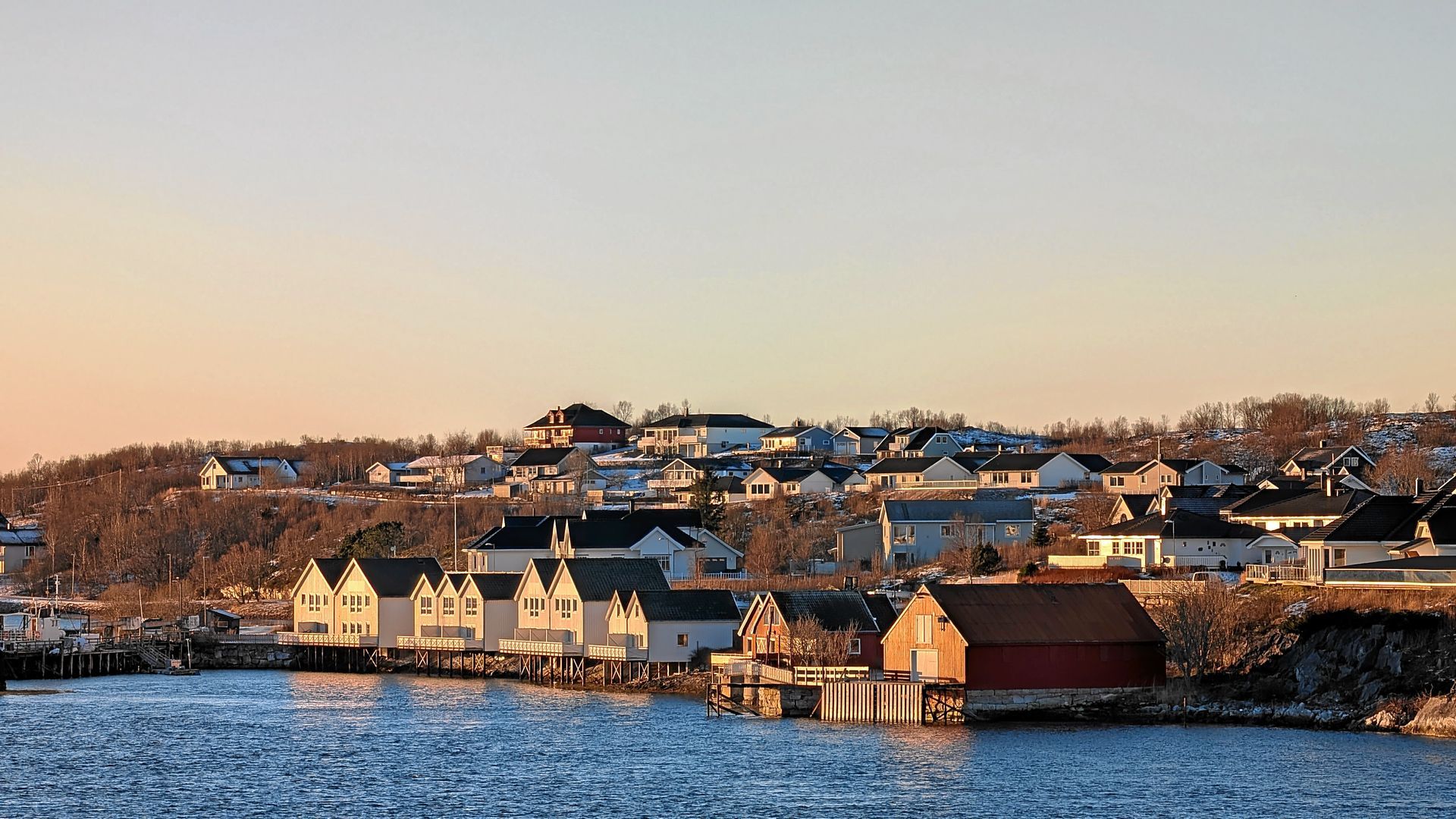 Küstenstadt bei Sonnenuntergang, weiße und rote Gebäude säumen das Wasser, orangefarbener Himmel.