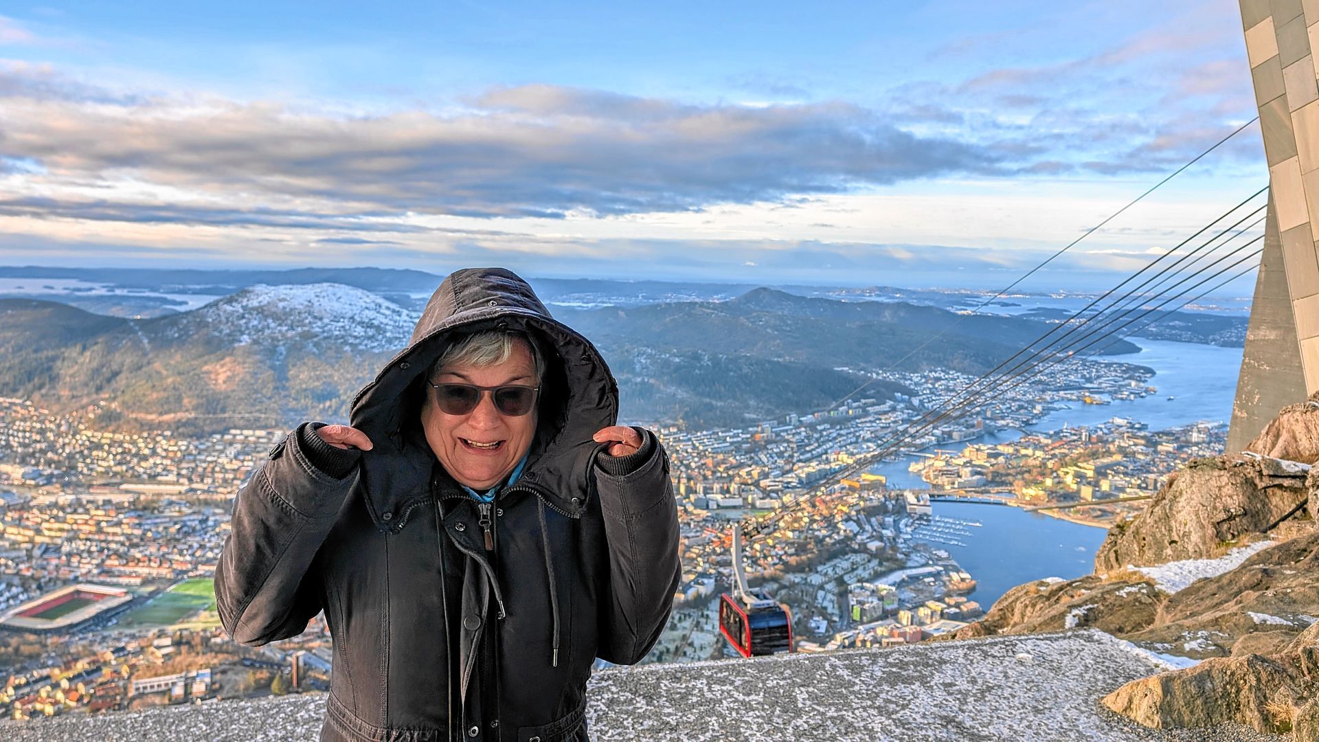 Eine Person in einem Kapuzenmantel lächelt auf einem schneebedeckten Berggipfel mit Blick auf eine Stadt und einen Hafen.
