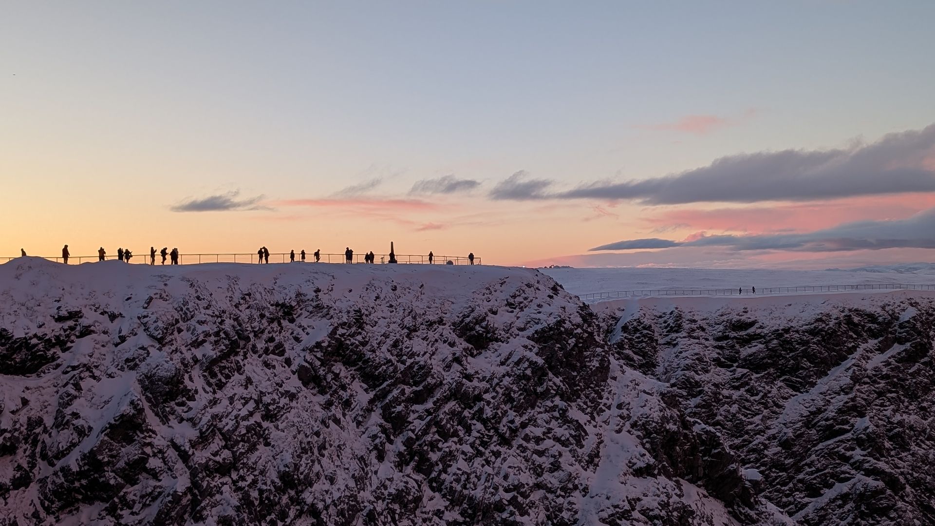 Schneebedeckter Bergkamm mit Menschen, deren Silhouetten sich vor einem pastellfarbenen Sonnenuntergang abzeichnen.