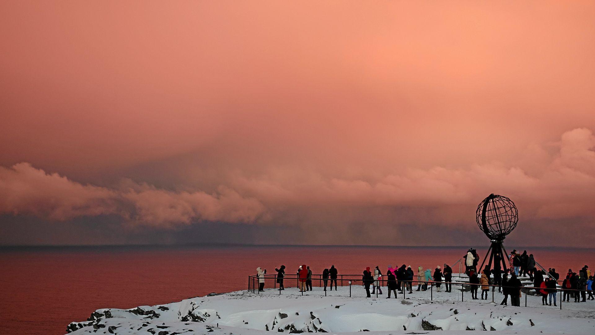Menschen in der Nähe eines Globus-Denkmals auf einer schneebedeckten Klippe, über dem Meer ein rosafarbener Himmel.