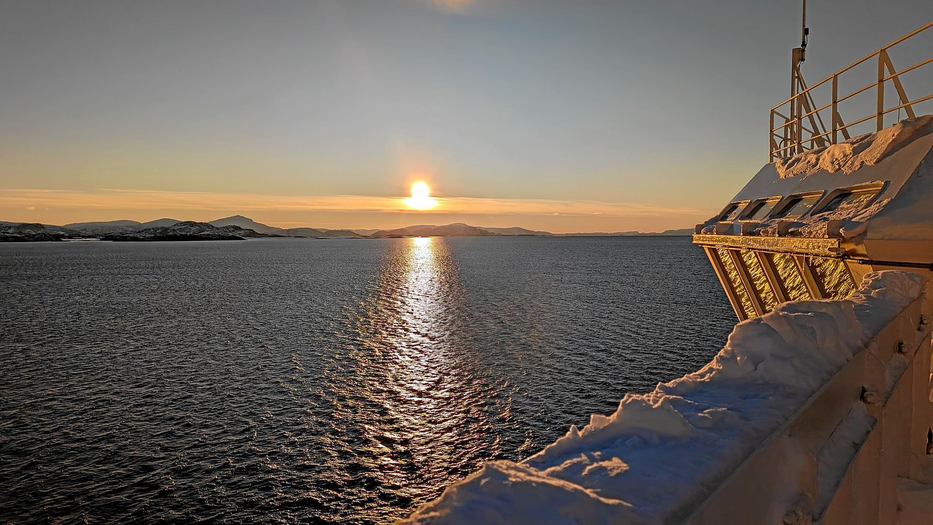 Sonnenuntergang über eisigem Wasser vom Deck eines Schiffes aus, mit Schnee, der ein reflektierendes goldenes Licht erzeugt.