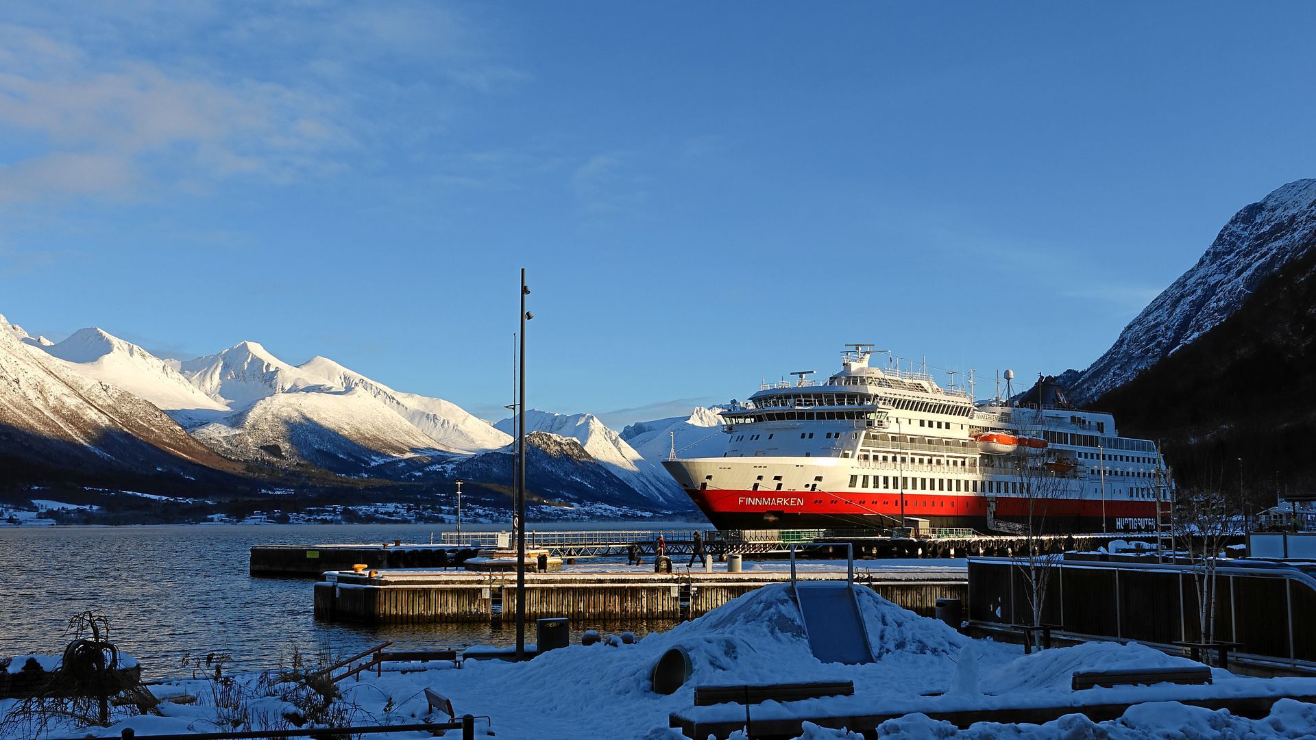 Ein Kreuzfahrtschiff liegt im verschneiten Hafen vor Anker, im Hintergrund die Berge, unter blauem Himmel.