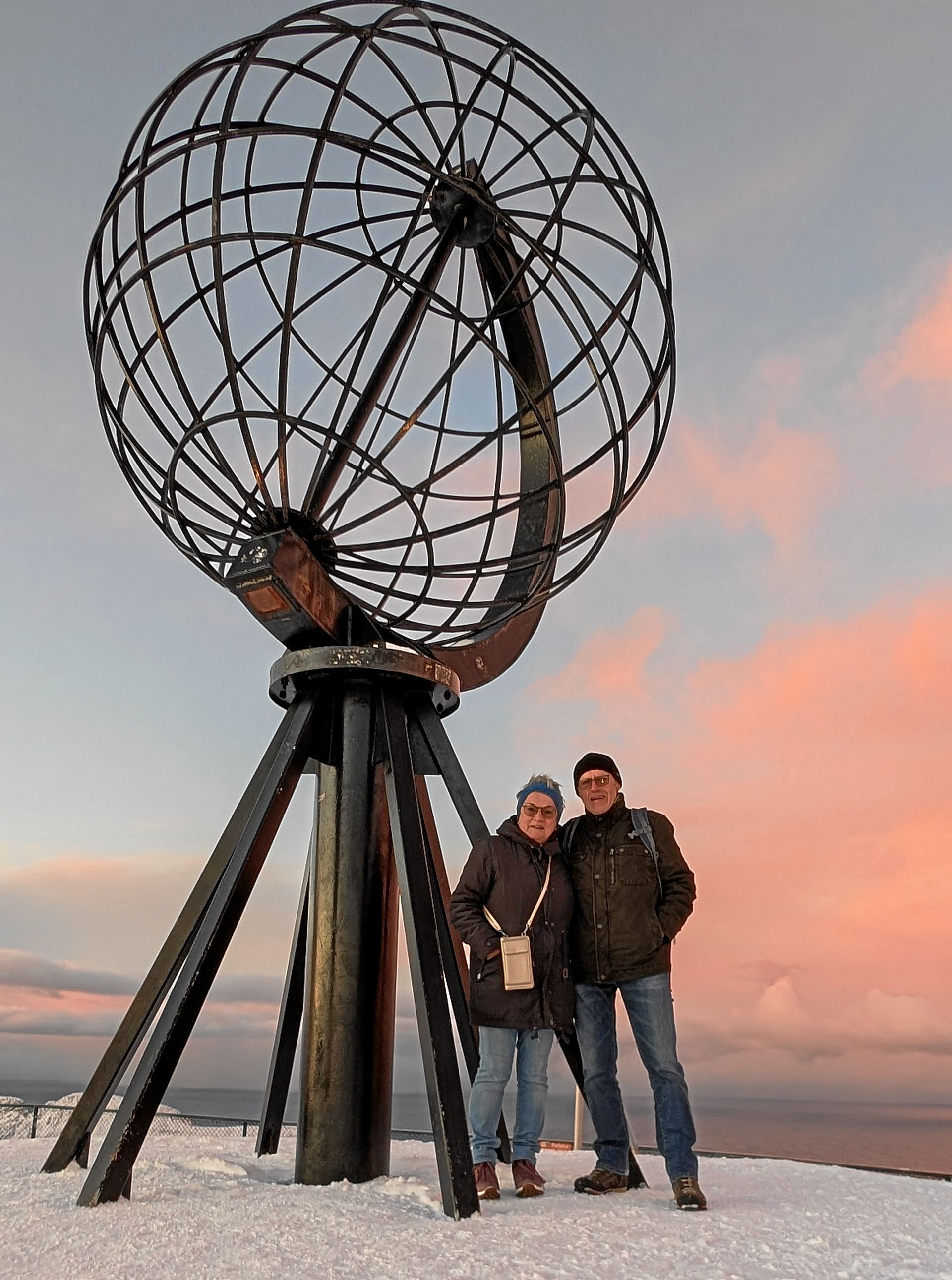Zwei Personen posieren vor einer großen Globus-Skulptur am Nordkap in Norwegen vor einem Sonnenuntergang.