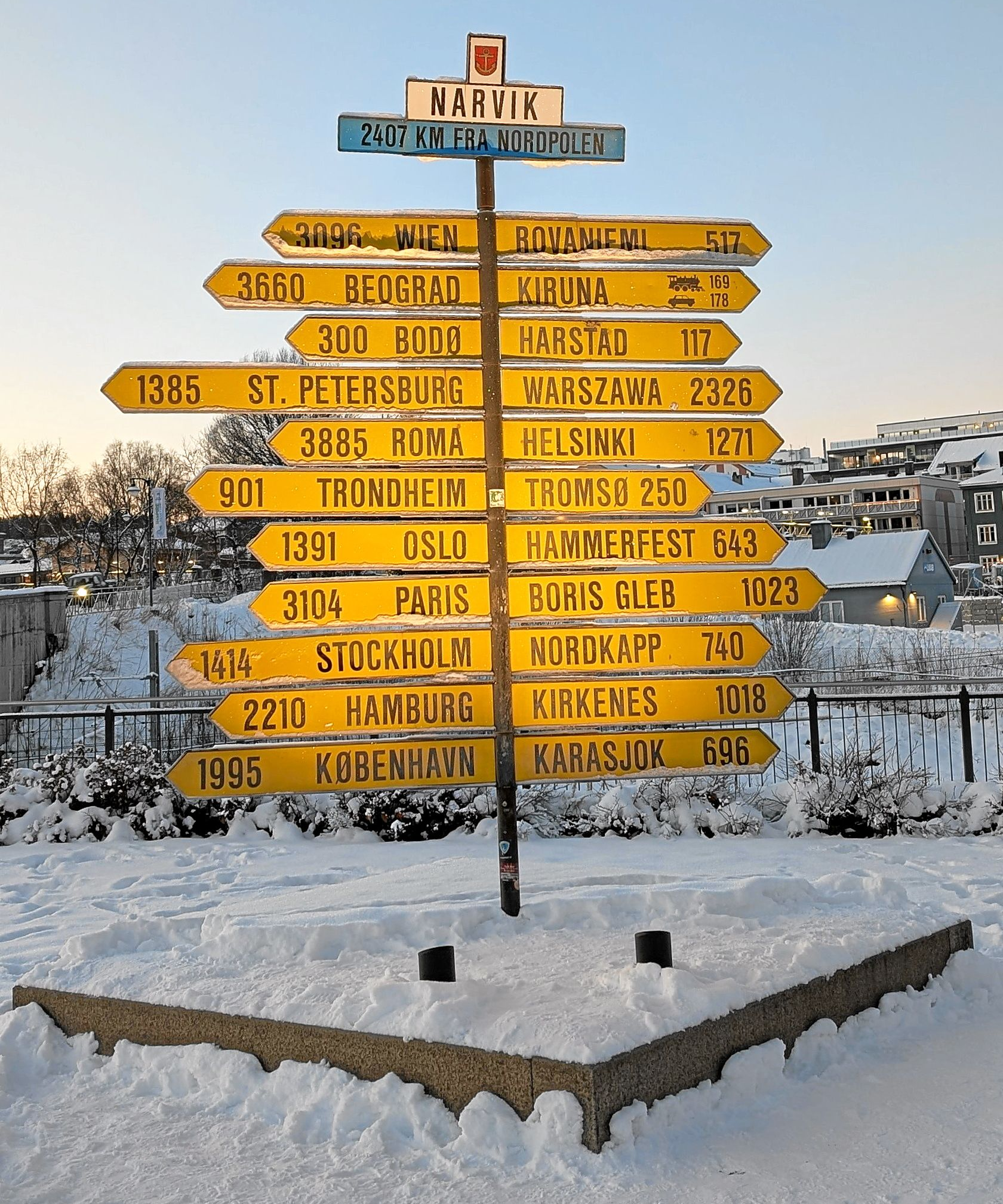Gelber Wegweiser im Schnee, der zu verschiedenen Städten mit Entfernungsangaben weist.