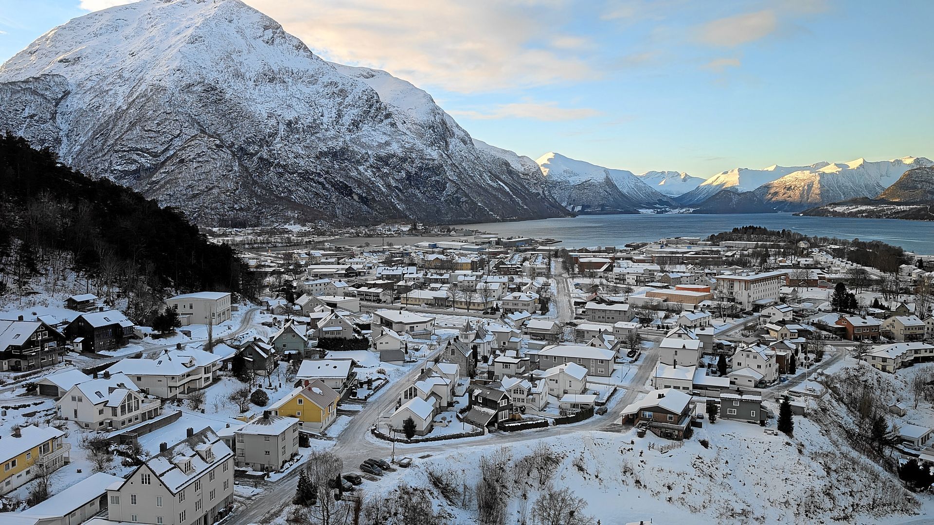 Eine schneebedeckte Stadt, eingebettet an einem Fjord, Berge im Hintergrund, blaues Wasser und ein strahlend blauer Himmel.