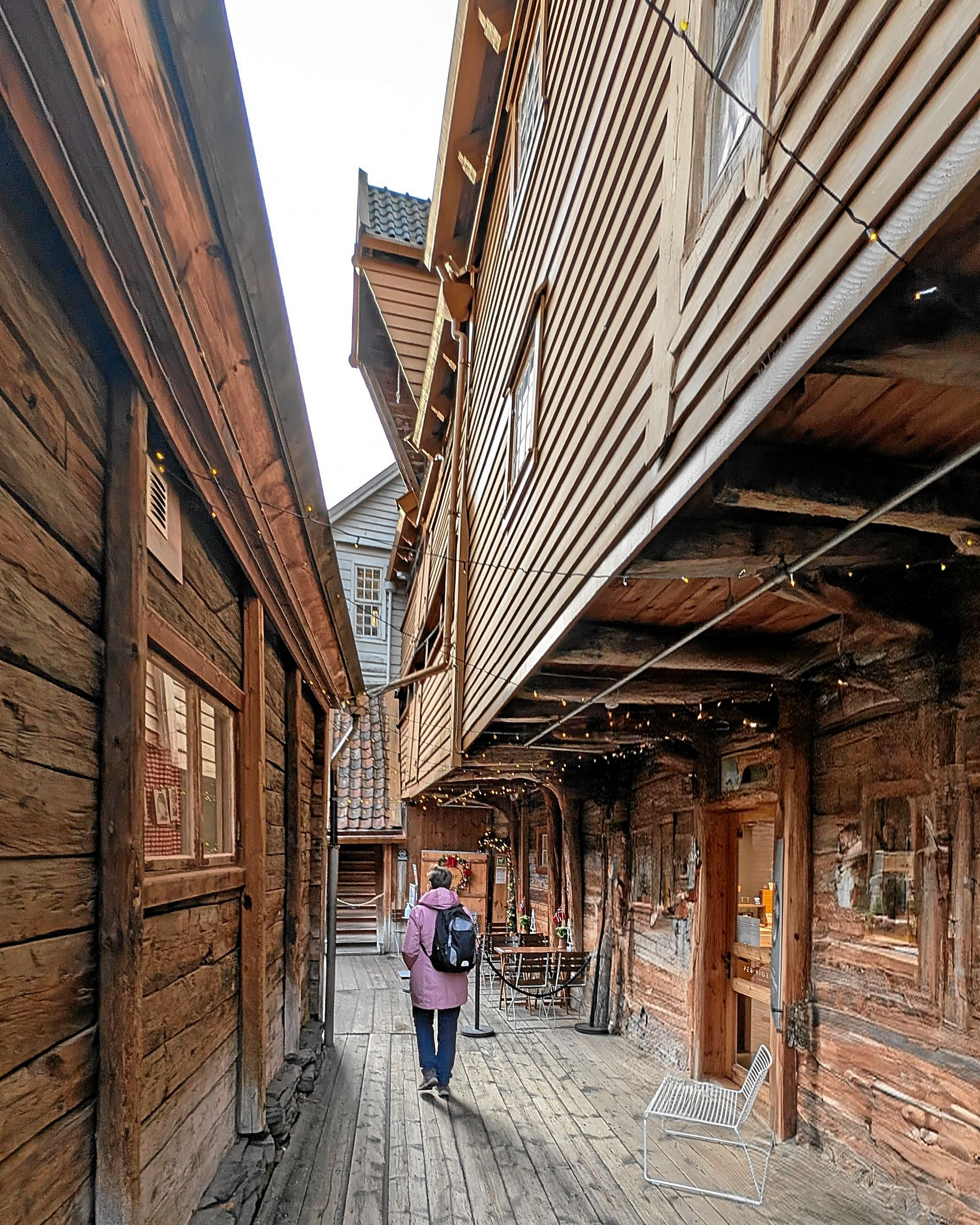 Schmale Holzgasse in Bergen, Norwegen. Eine Person geht auf die Kamera zu. Braune Gebäude, bedeckter Himmel.