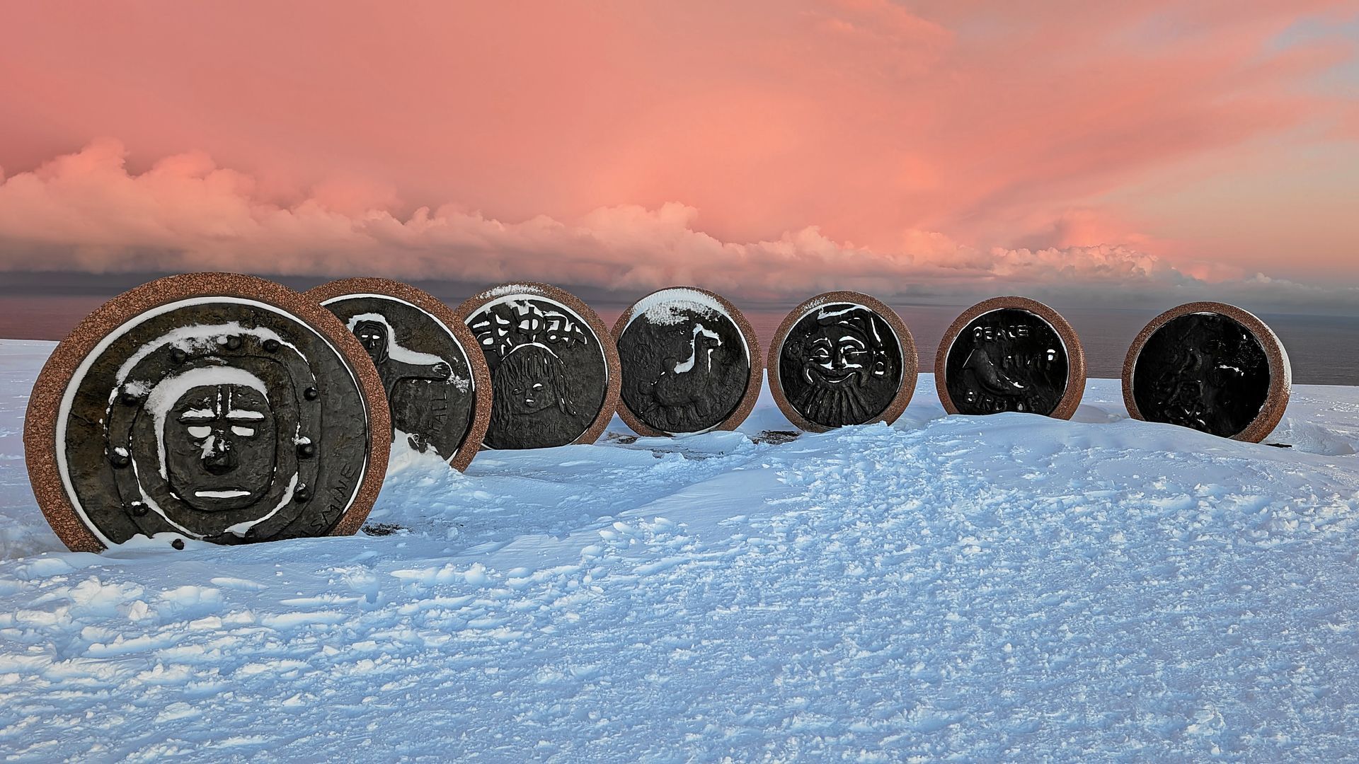 Sieben kreisförmige, aus Stein gehauene Skulpturen im Schnee; im Hintergrund rosa Himmel.