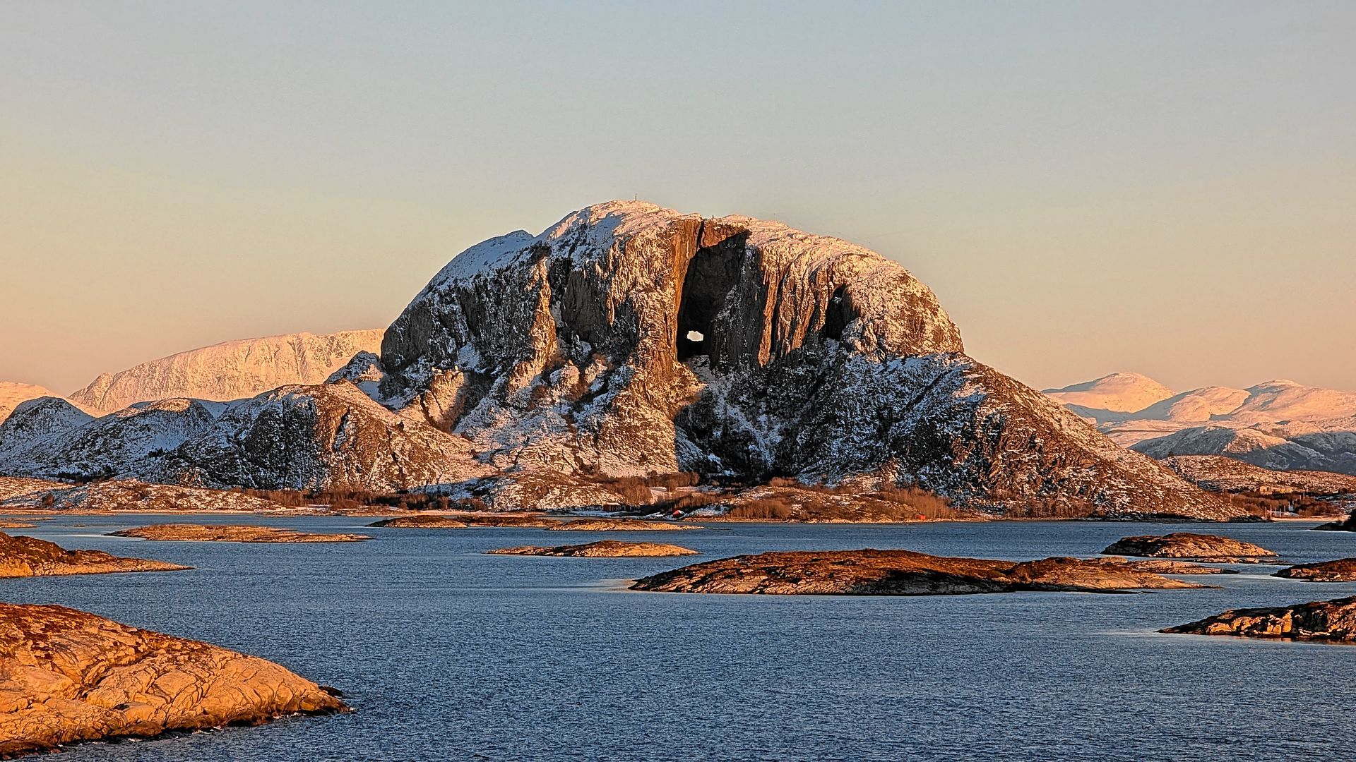 Ein schneebedeckter Berg mit einem Loch darin, umgeben von Wasser und kleinen Inseln, unter einem goldenen Sonnenuntergang.