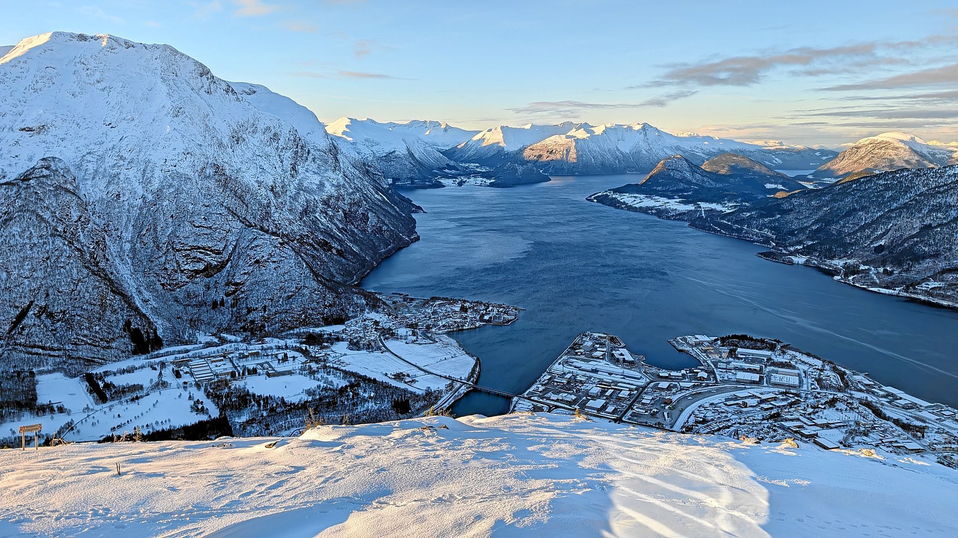 Schneebedeckte Berge überragen einen Fjord, an dessen Ufer sich eine Stadt schmiegt.