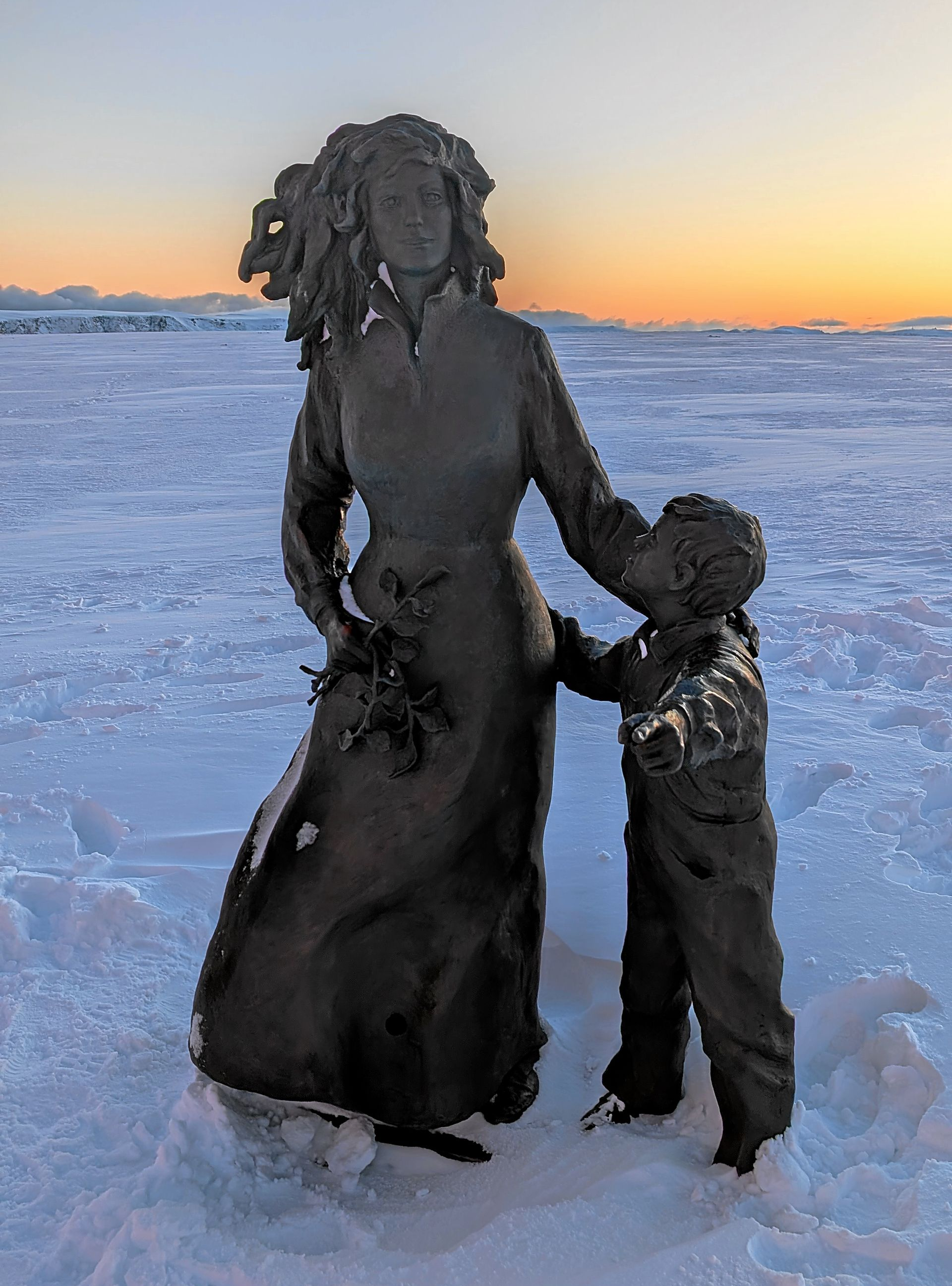 Skulptur einer Frau mit Kind im Schnee; Sonnenuntergang im Hintergrund.