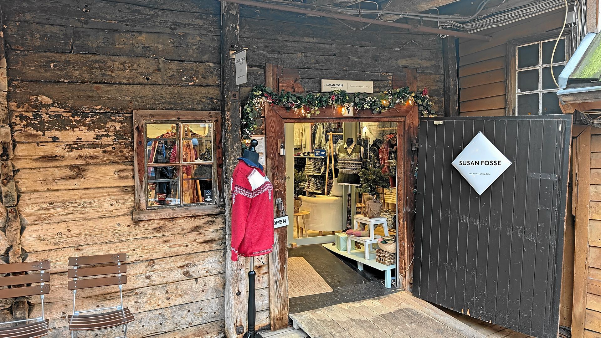 Außenansicht eines Holzladens mit offener Tür, in dem Waren ausgestellt sind, und einem Schild.