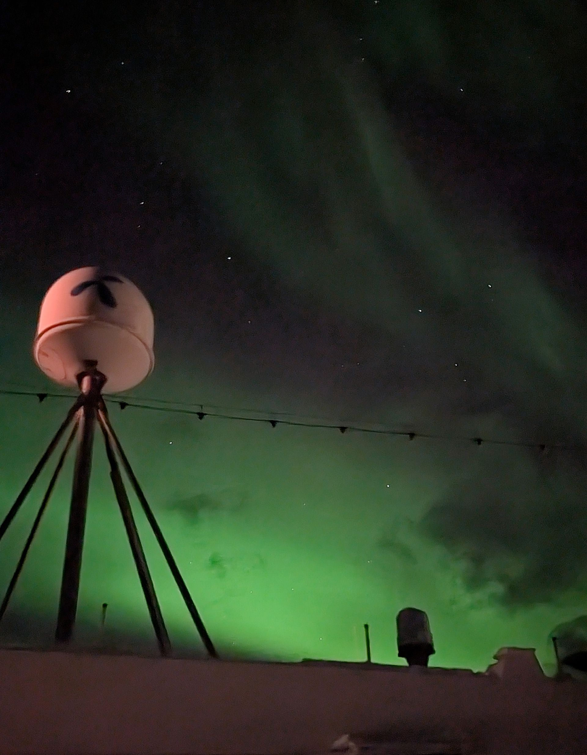 Die Aurora borealis erleuchtet den Nachthimmel. Weiße Geräte stehen auf einem Dach; Sterne sind sichtbar.
