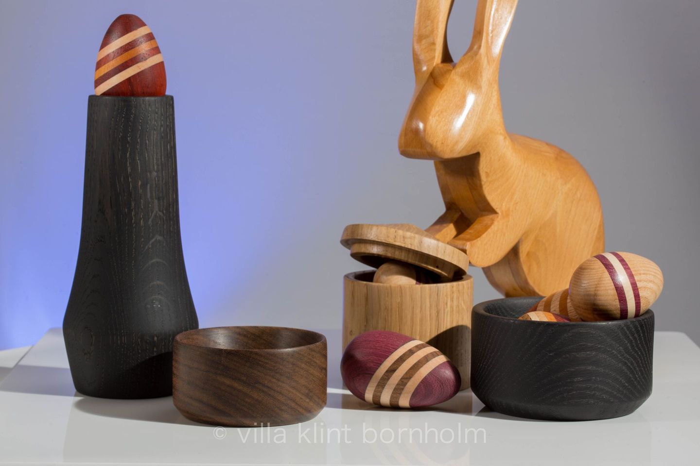 Osterbild: ein Osterhase versteckt mehrfarbig gestreifte Ostereier in Holzdosen und Vasen