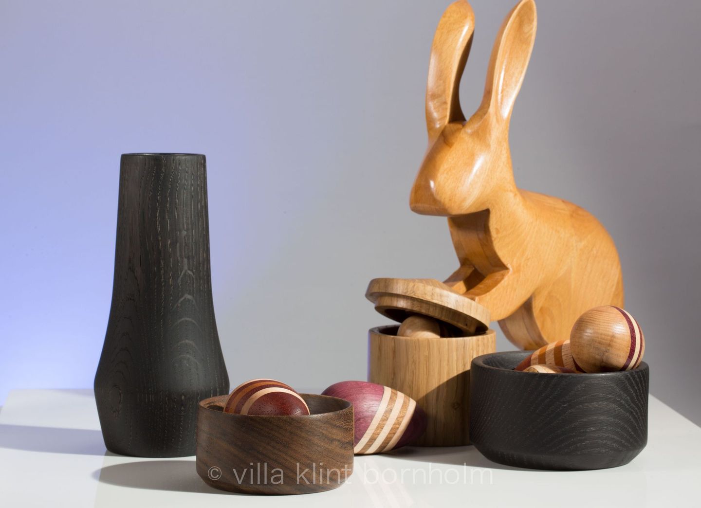 Osterbild: ein Osterhase versteckt mehrfarbig gestreifte Ostereier in Holzdosen