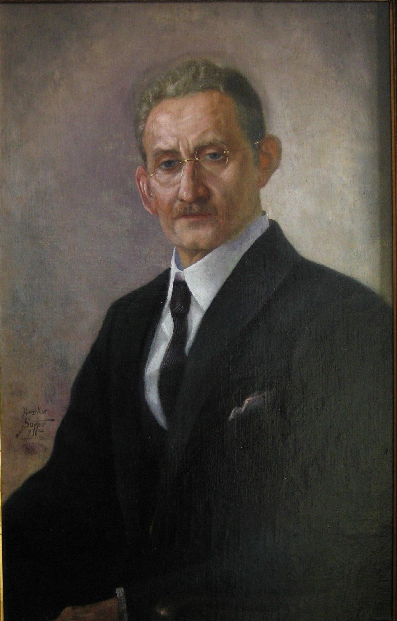 Porträt eines Mannes im Anzug mit Brille und ernstem Gesichtsausdruck vor neutralem Hintergrund.