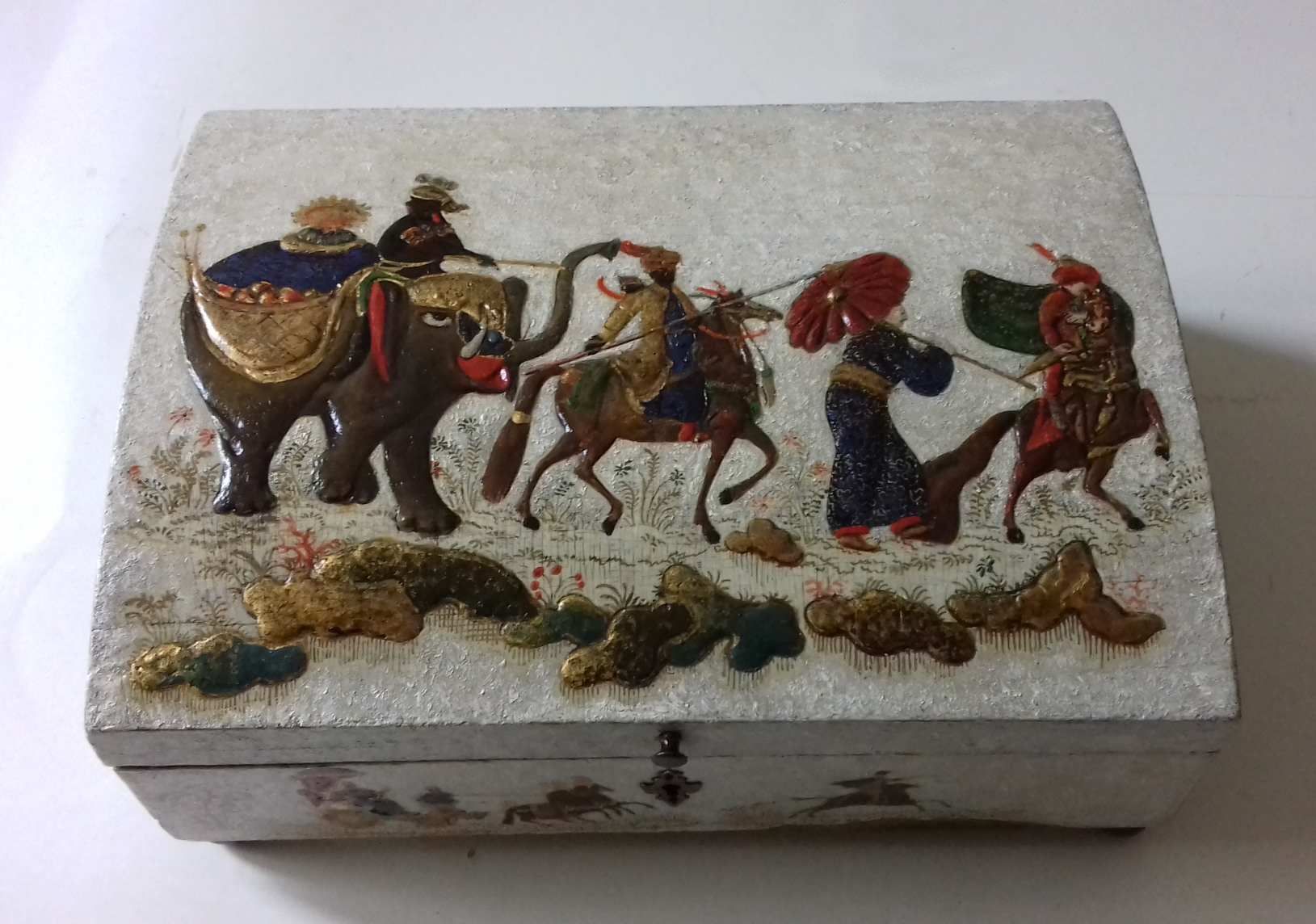 Dekorative Schachtel mit Elefantenmotiv und Figuren in einer Parade. Gold- und Juwelentöne auf weißem, strukturiertem Hintergrund.