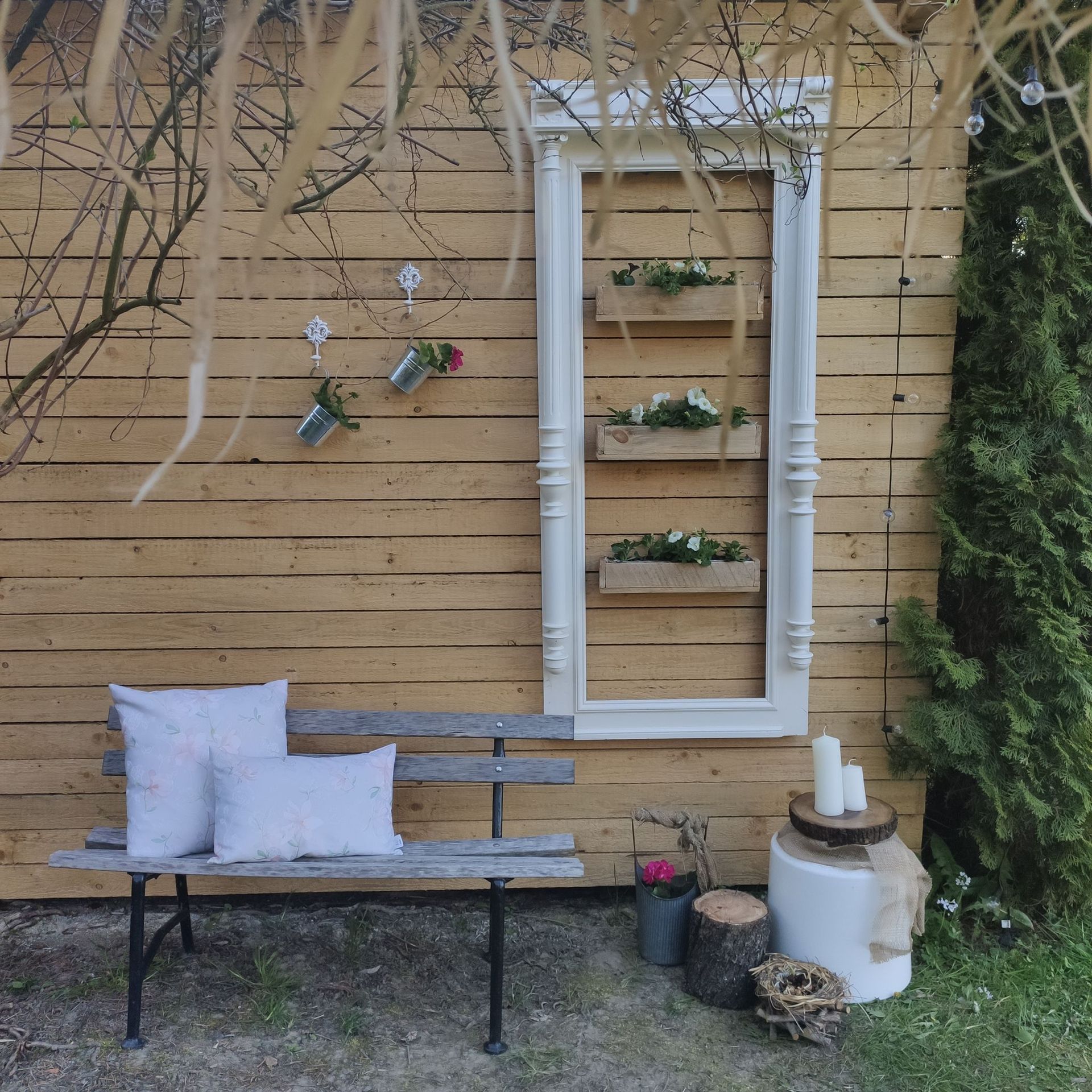 Gemütliche Gartenterrasse mit weißer Bank, Kissen, Wandpflanzgefäßen und dekorativen Laternen vor einem Holzzaun