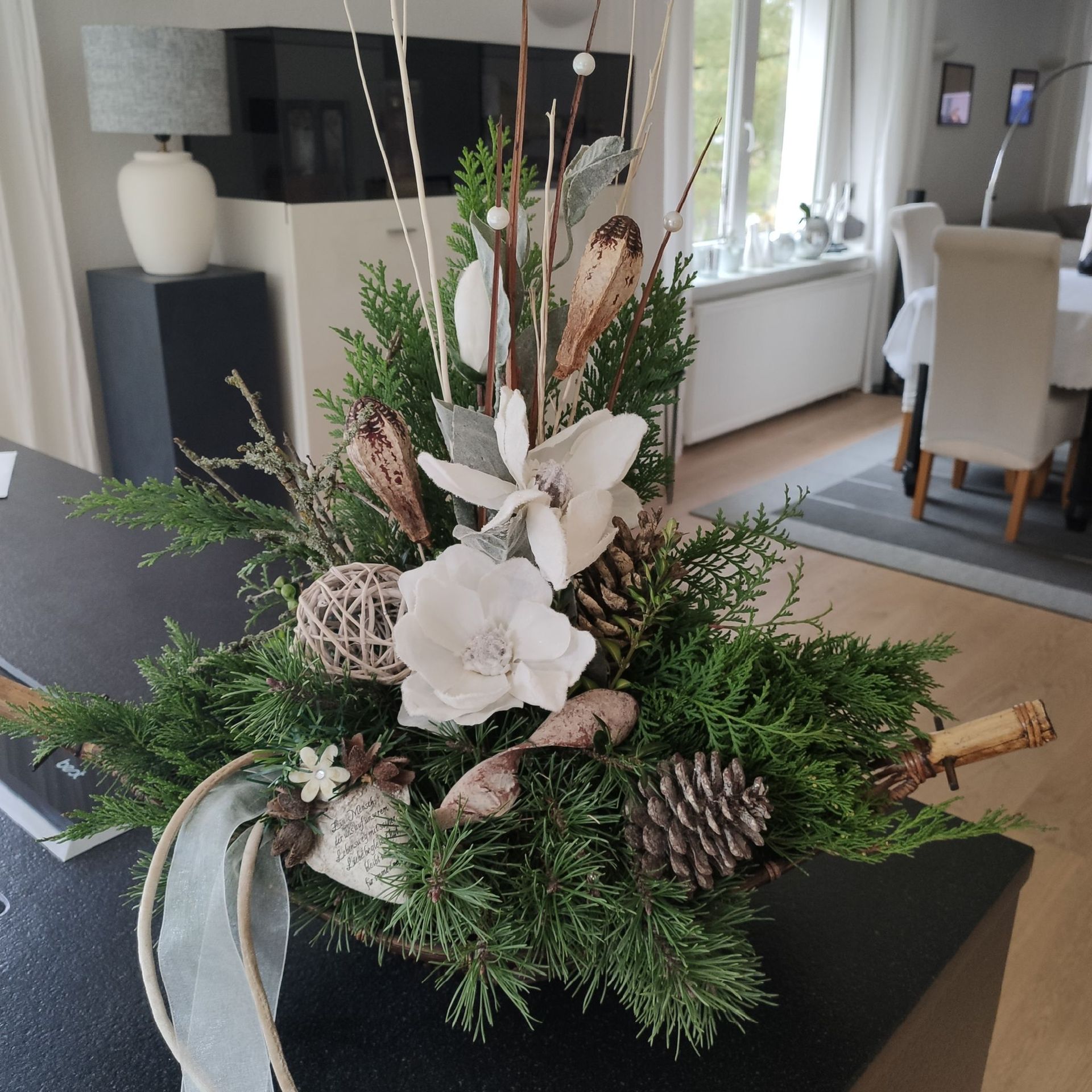 Festliches Blumenarrangement mit Tannenzapfen, weißen Blüten und Grün auf einem schwarzen Tisch
