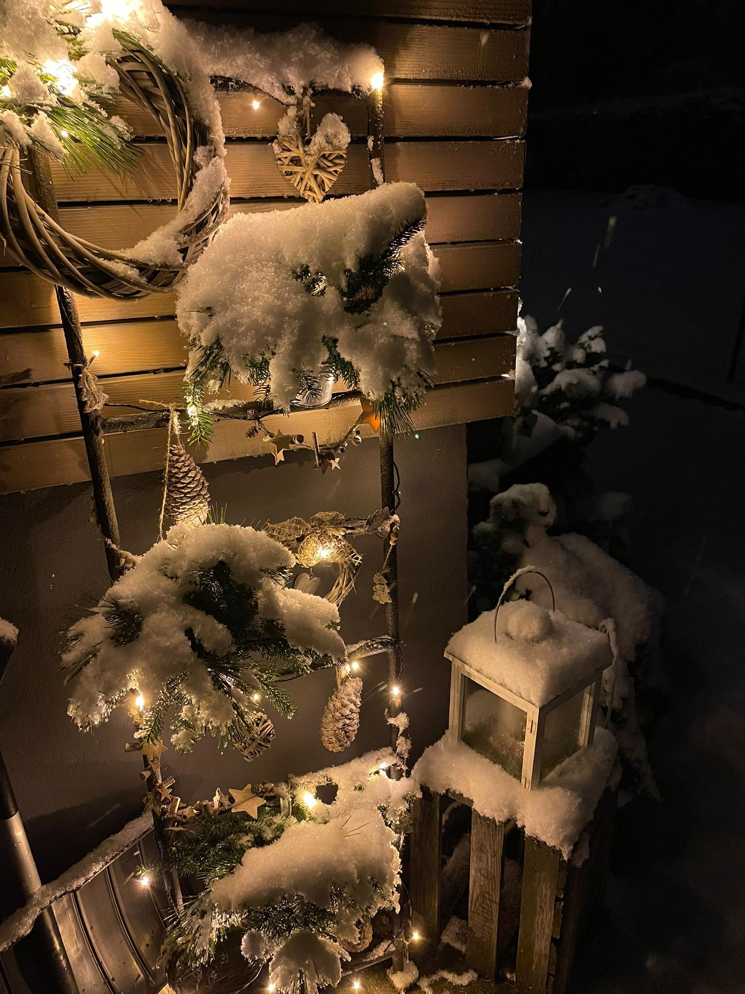 Eine schneebedeckte Veranda mit Lichterketten, Kränzen und einer Laterne bei Nacht