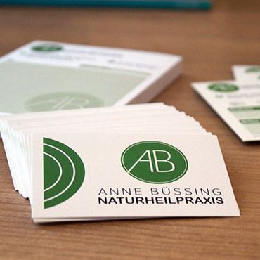 Visitenkarten auf einem braunen Tisch. Die Karten zeigen das runde grüne Logo mit den Initialien AB. Darunter steht Anne Büssing Naturheilpraxis. im Hintergrund liegt unscharf der Rezeptblock und die Terminkarten.