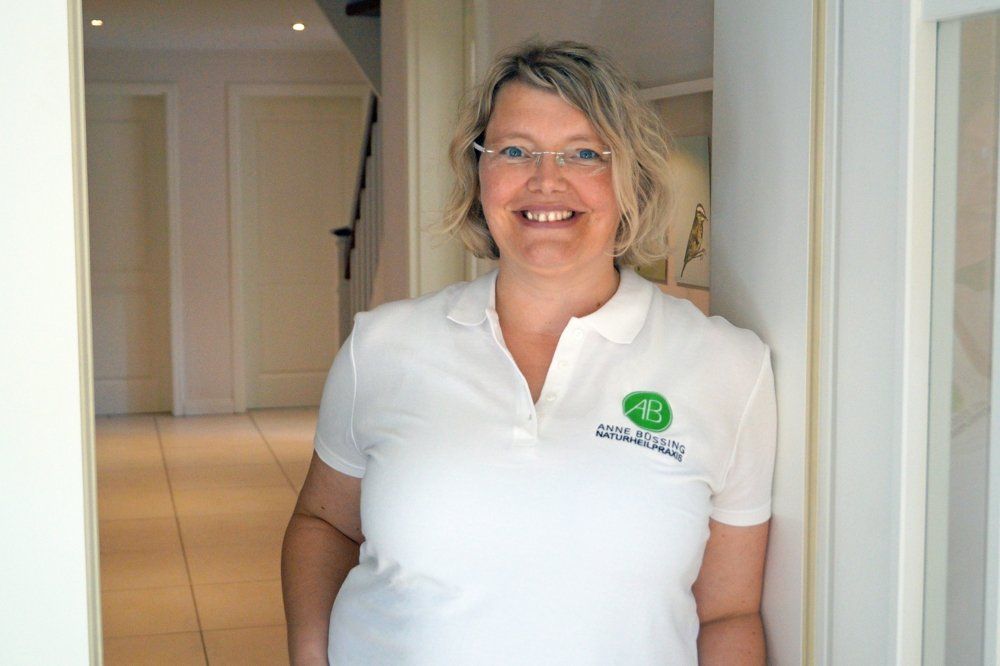 Anne Büssing in den Türrahmen gelehnt. Sie hat ein weißes Poloshirt mit Logo der Naturheilpraxis auf der Linken Brustseite gestickt. Im Hintergrund sind ein heller Flur mit zwei Türen und einer Treppe zu sehen.