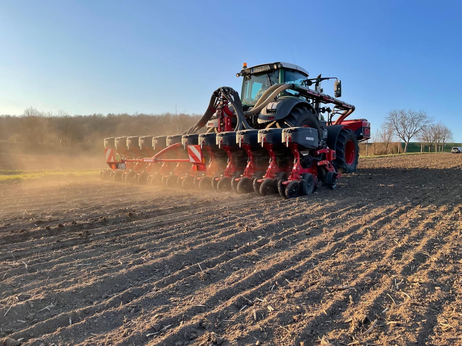 Fendt-Traktor mit 12-reihigem Horsch-Einzelkornsägerät bei der Aussat auf einem frisch bearbeiteten Acker, bei blauem Himmel.