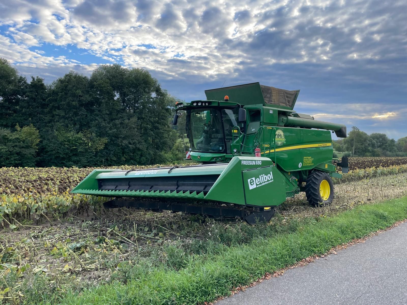 John Deere-Mähdrescher beim dreschen eines Sonneblumenfeldes, im Hintergrund grüner Wald unter bewölktem Himmel.