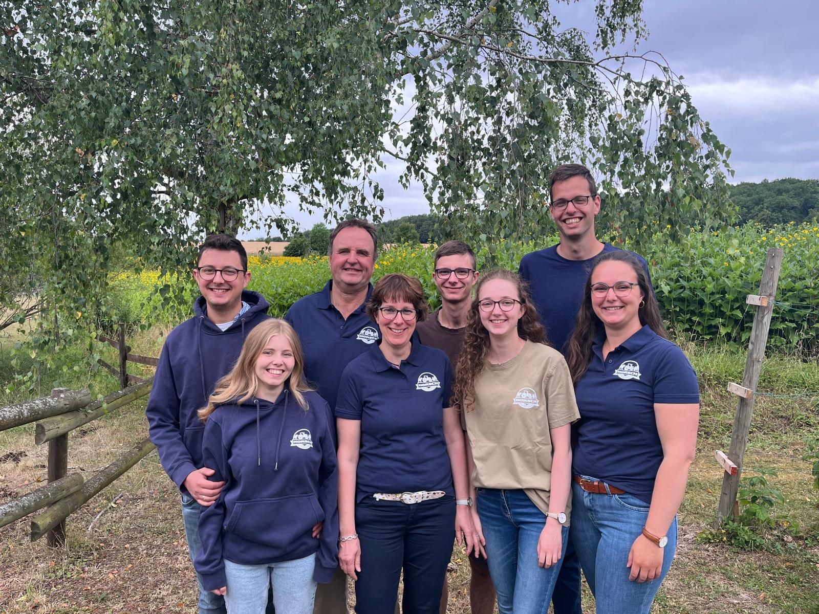Gruppenfoto von acht Peronen in Arbeitskleidung des Hofs. Im Hintergrund ländliche Kulisse mit Bäumen und Feldern.