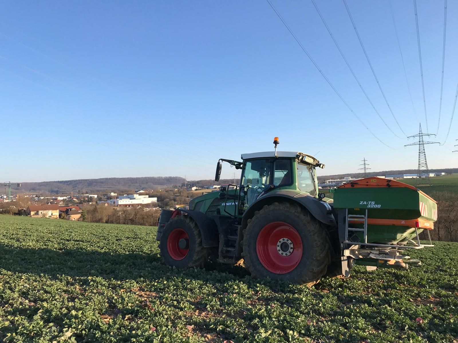 Fendt-Trakto mit Amazone-Düngerstreuer auf einem bepflanzten Feld, im Hintergrund ein Industriegebiet unter blauem Himmel.