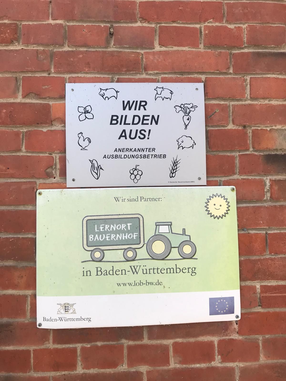 Schilder Wir bildem aus - Anerkannter Ausbildungsbetrieb
und Lernort Bauernhof in Baden-Württemberg, an einer Backsteinwand.