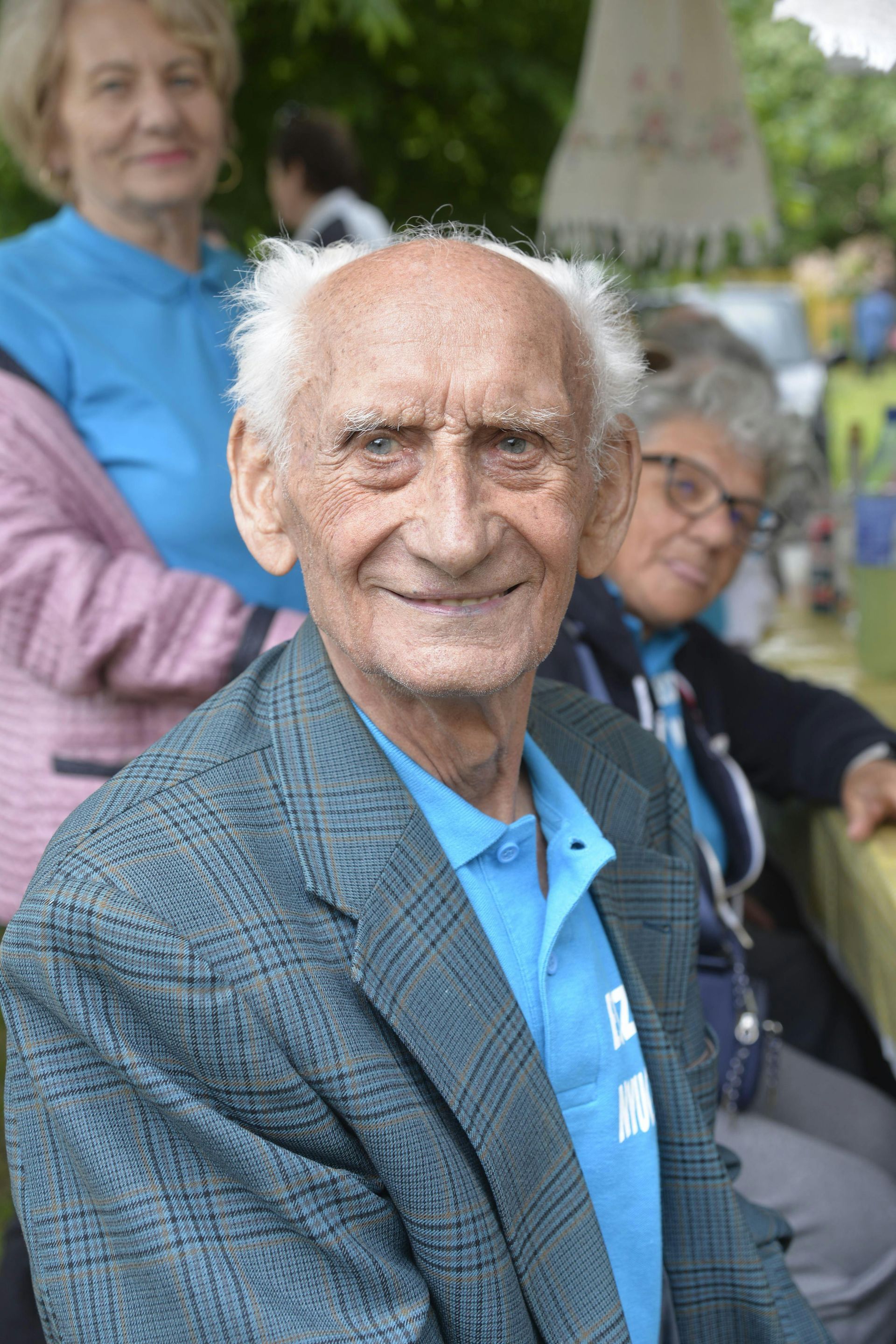 Älterer Mann in kariertem Sakko lächelt in die Kamera, Frau im Hintergrund