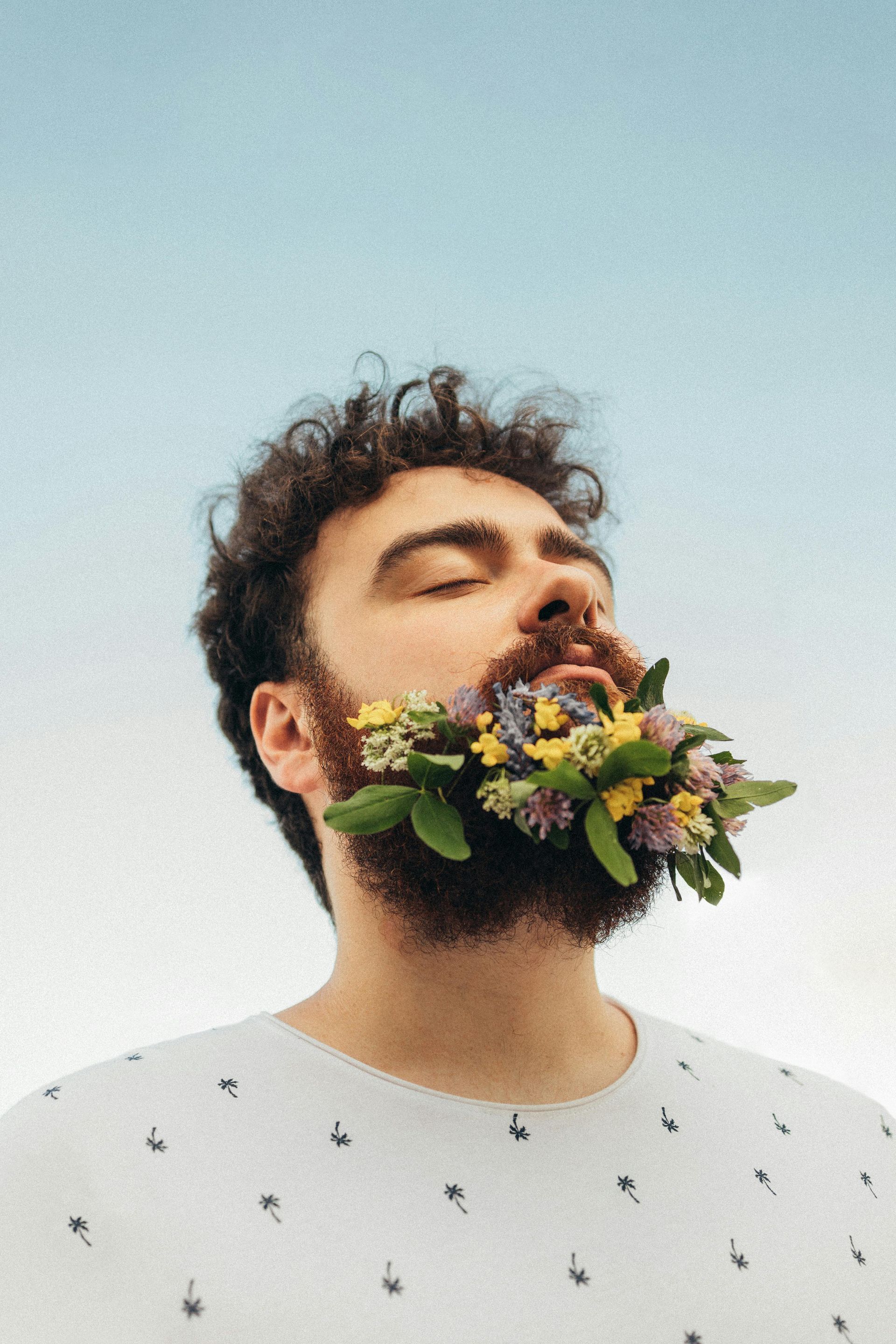 Mann mit geschlossenen Augen und Blumen im Bart vor Himmel