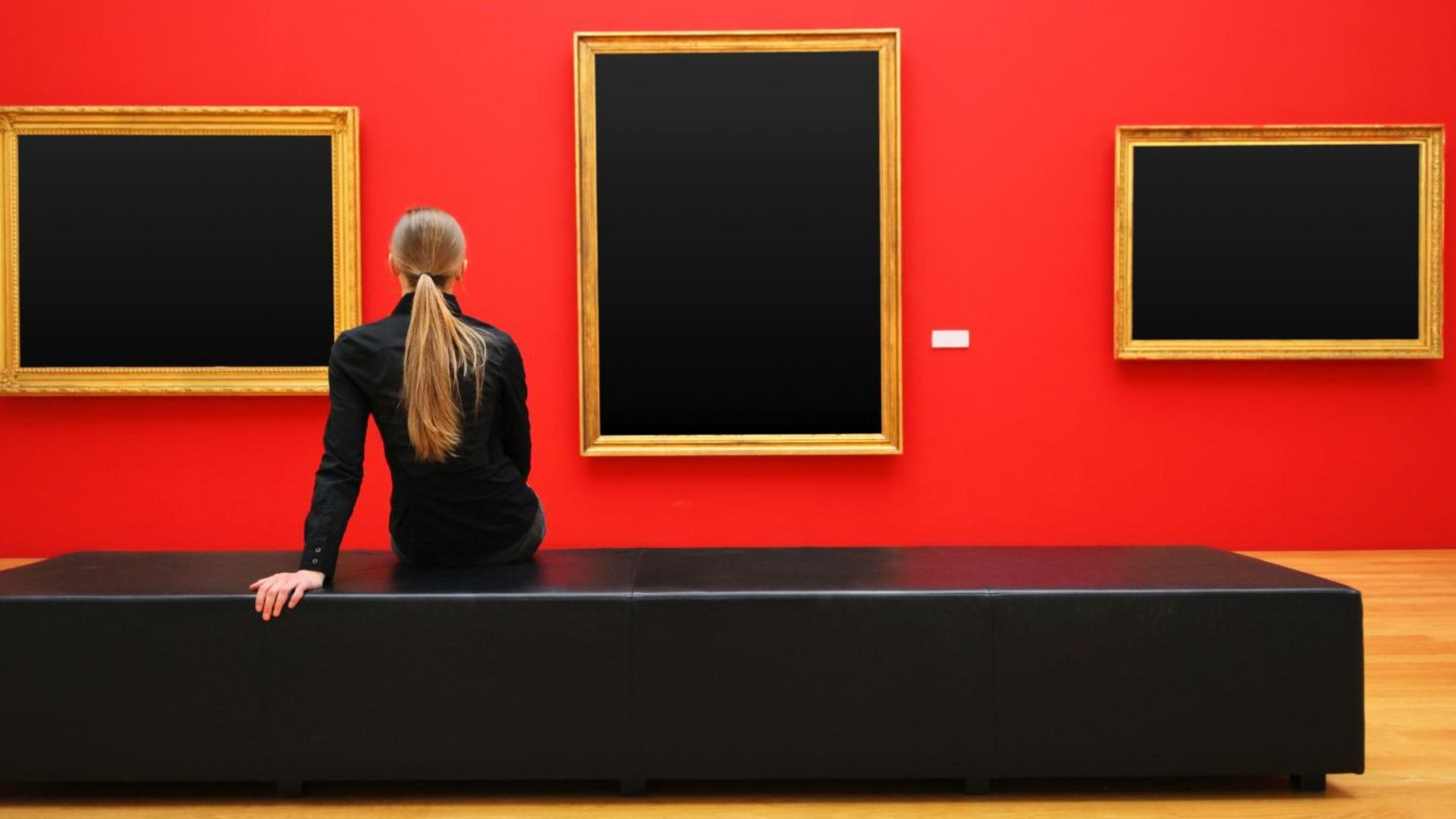 Entdeckt die Kunst Eine Frau sitzt auf einer Bank und betrachtet ein leeres, gerahmtes Bild an einer roten Wand.