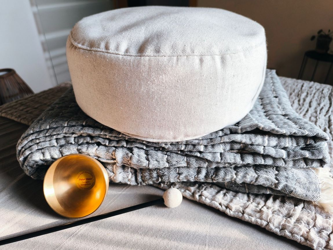 Ein Yogakissen liegt auf einer gefalteten Decke, eine kleine Klangschale steht im Vordergrund.