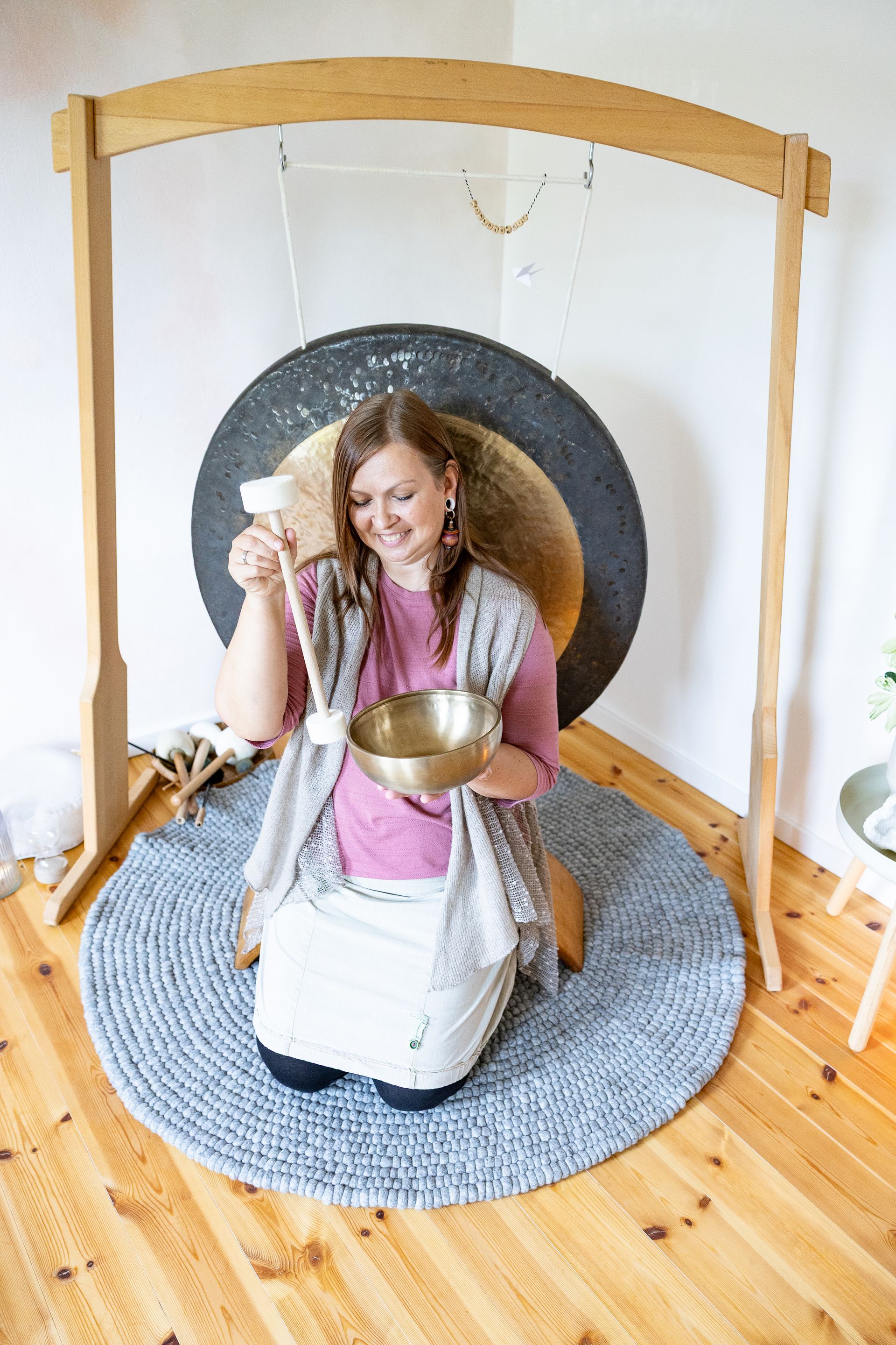 Julia Pantou sitzt mit dem Rücken vor einem Gong, lächelt und hält eine Klangschale in der Hand. Diese wird von ihr angetönt.