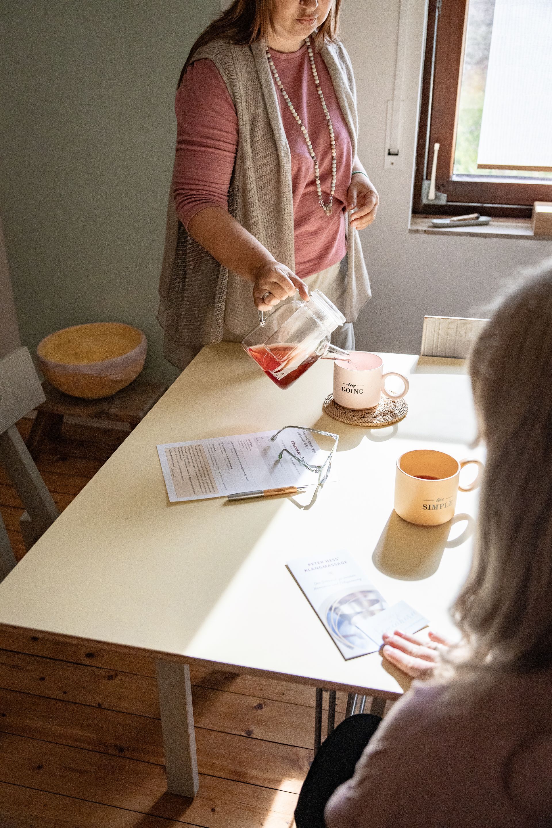 Julia Pantou schenkt Tee in eine Tasse ein, eine zweite Frau sitzt davor. Beide befinden sich am Tisch, im Kreativraum.