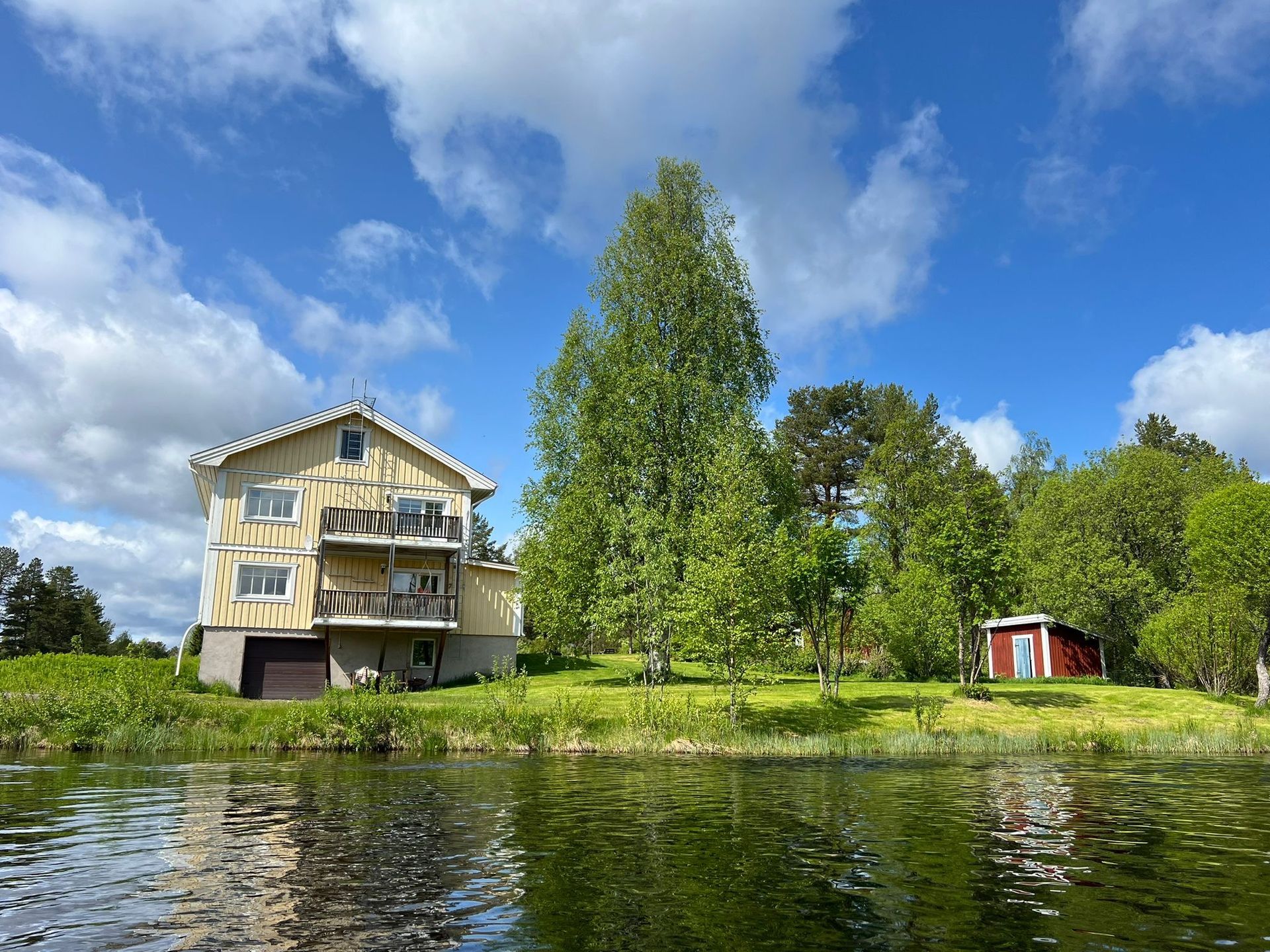 Zweistöckiges Haus mit Balkon an einem Seeufer, unter einem bewölkten blauen Himmel. Roter Schuppen im Hintergrund.