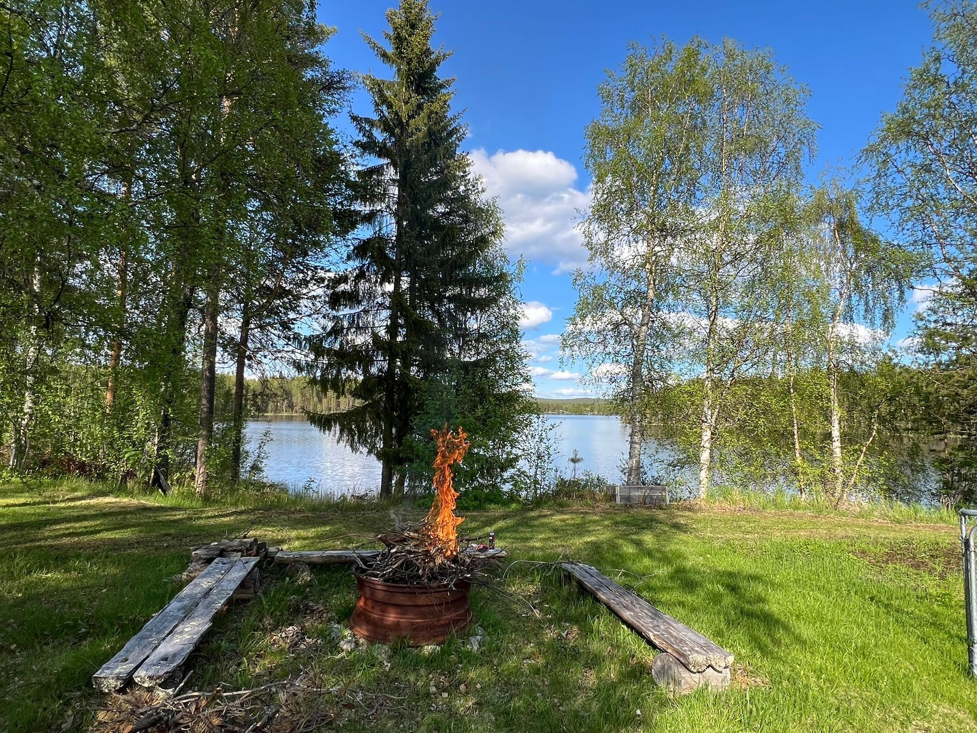 Lagerfeuer am See. Grünes Gras und Bäume rahmen die Szene unter einem blauen Himmel ein. Villa Aurora Moskosel