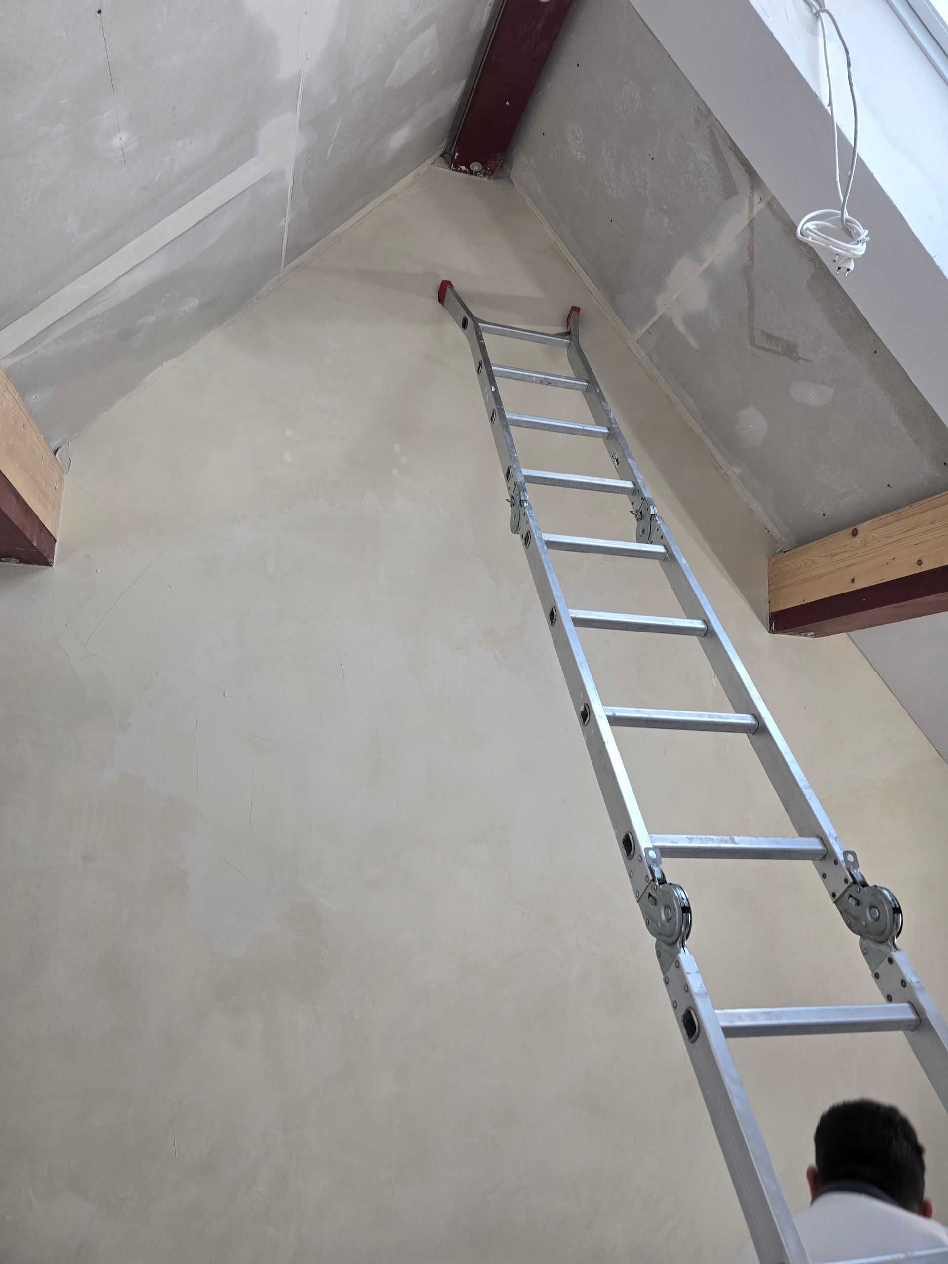 Eine Leiter lehnt an einer hellen Wand und reicht bis zur schrägen Decke. Am Fuß der Leiter befindet sich eine Person.