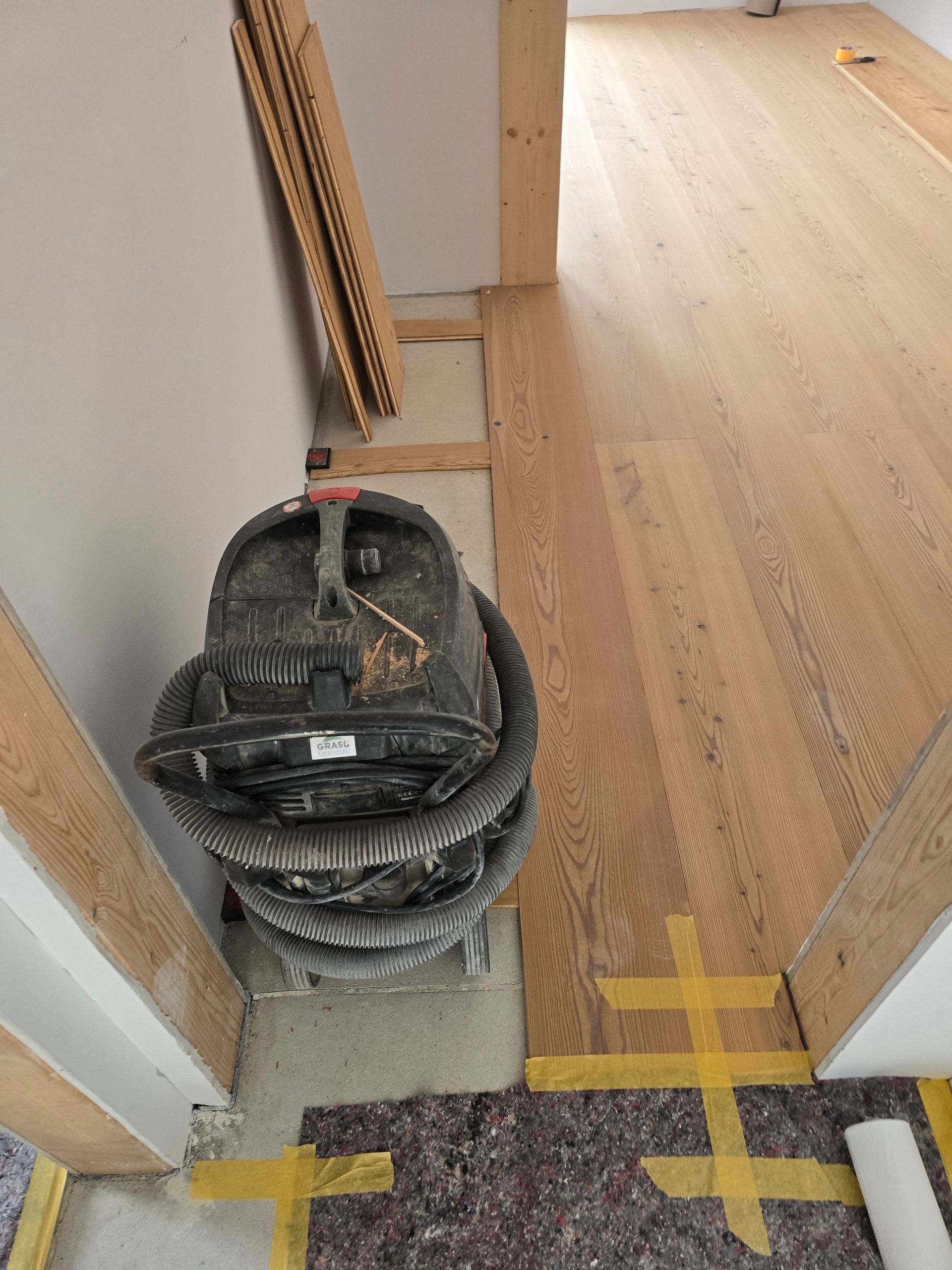 Staubsauger im Türrahmen eines Zimmers mit unfertigem Holzboden; Bauarbeiten im Gange.